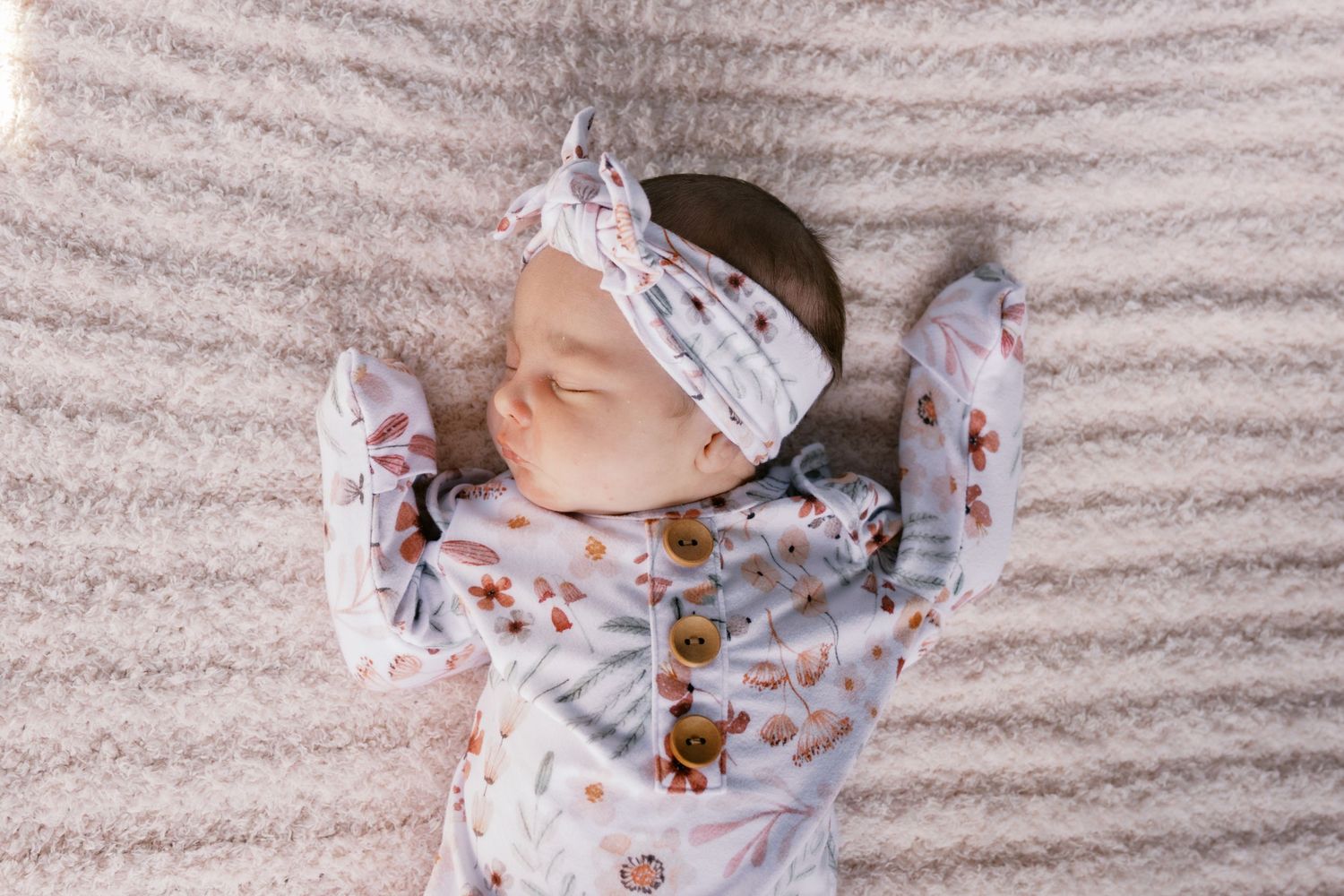 Newborn baby sleeping peacefully in floral outfit and matching knotted headband on soft white blanket.