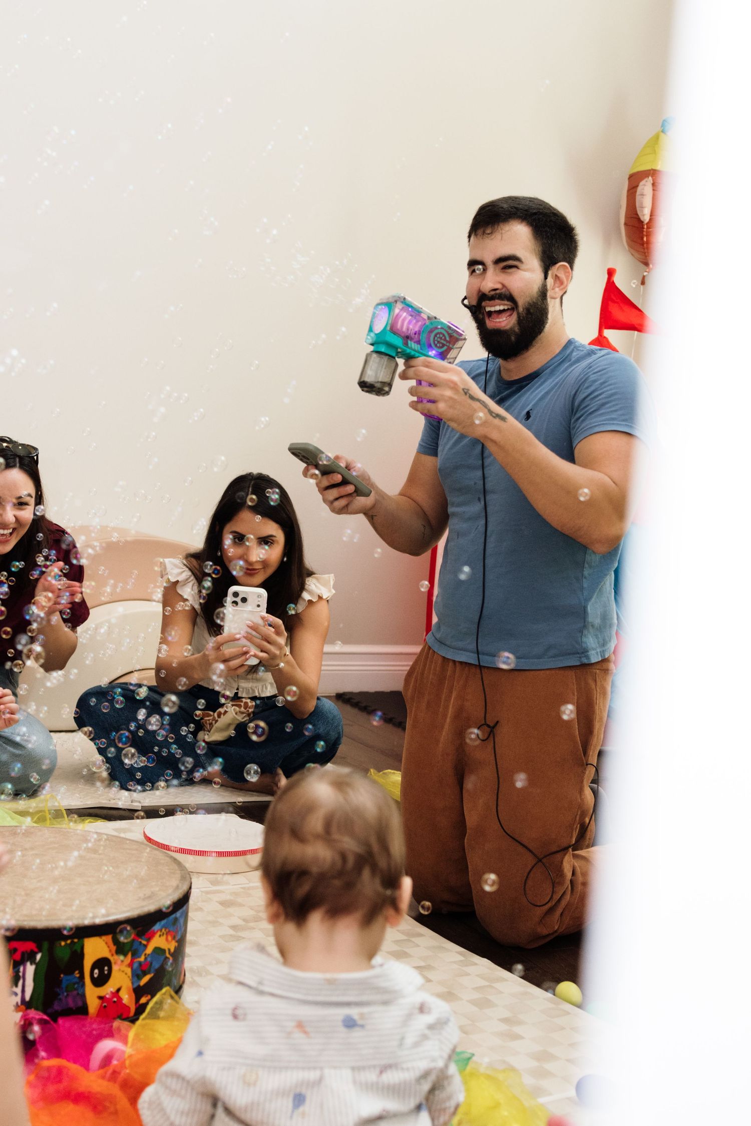 Man shoots bubble gun at baby during colorful indoor birthday party while guests sit on floor taking photos.