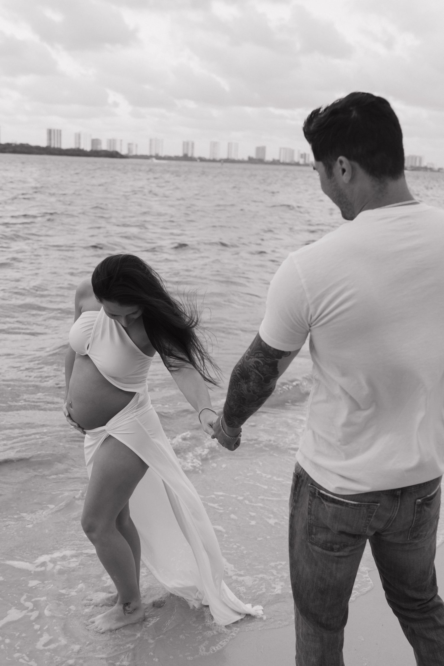 Black and white beach maternity photo of pregnant woman in white dress holding hands with tattooed man by the ocean.