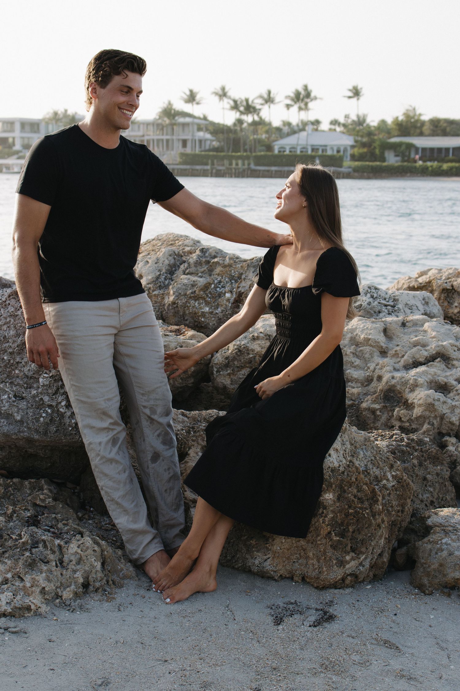 A couple shares a romantic moment on a rocky beach with coastal homes in the background.