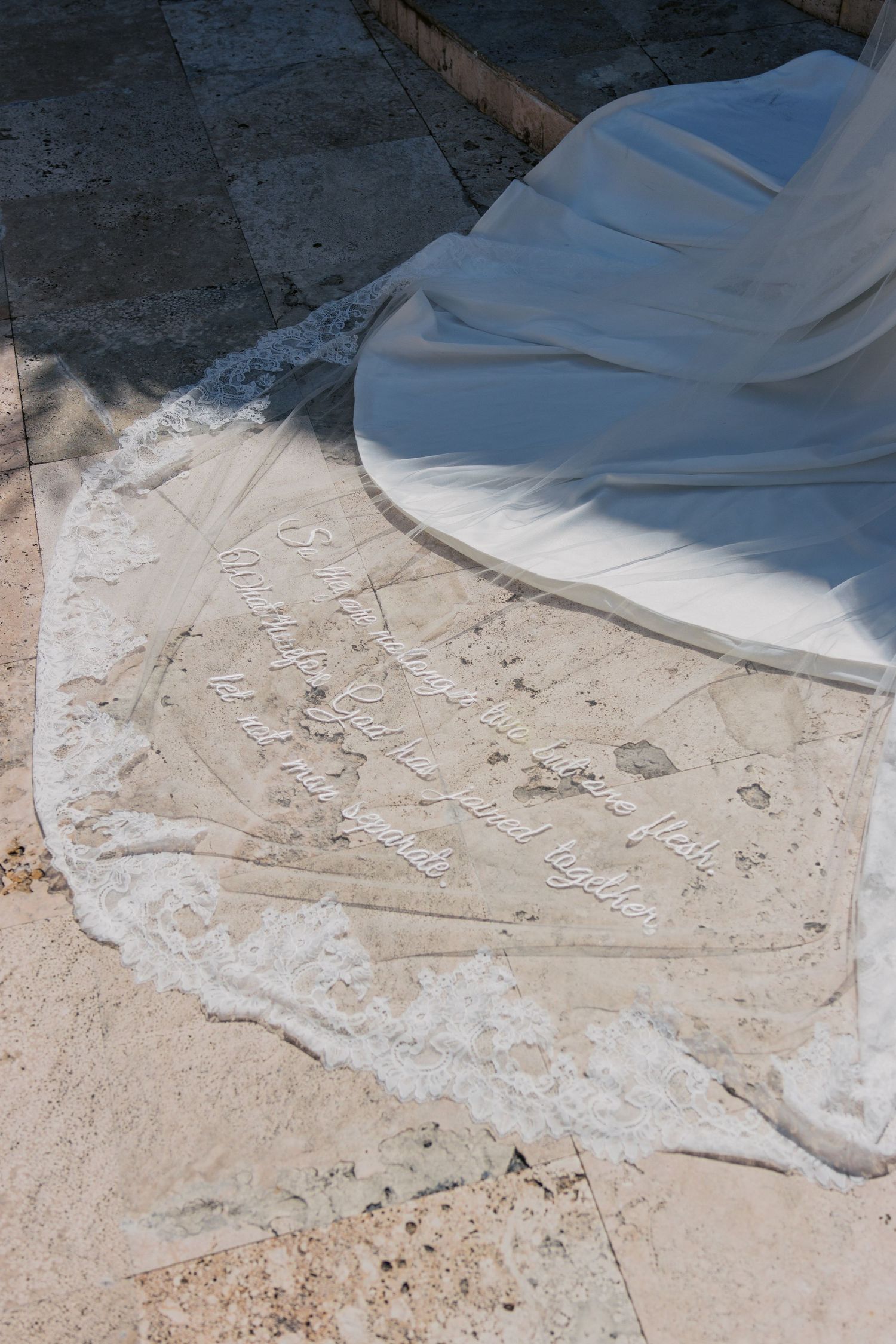 Wedding dress train with lace trim spread on stone pavement, featuring embroidered script text in sunlight.