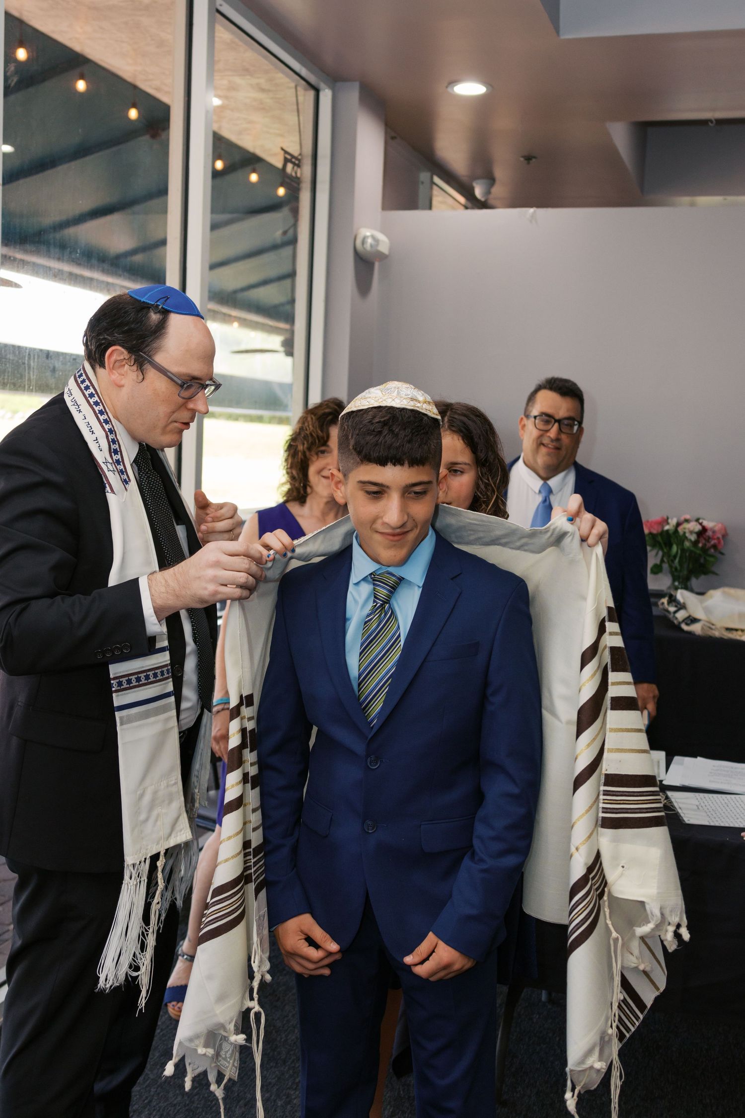 Young boy in blue suit receives tallit during Bar Mitzvah ceremony, surrounded by family in formal attire.