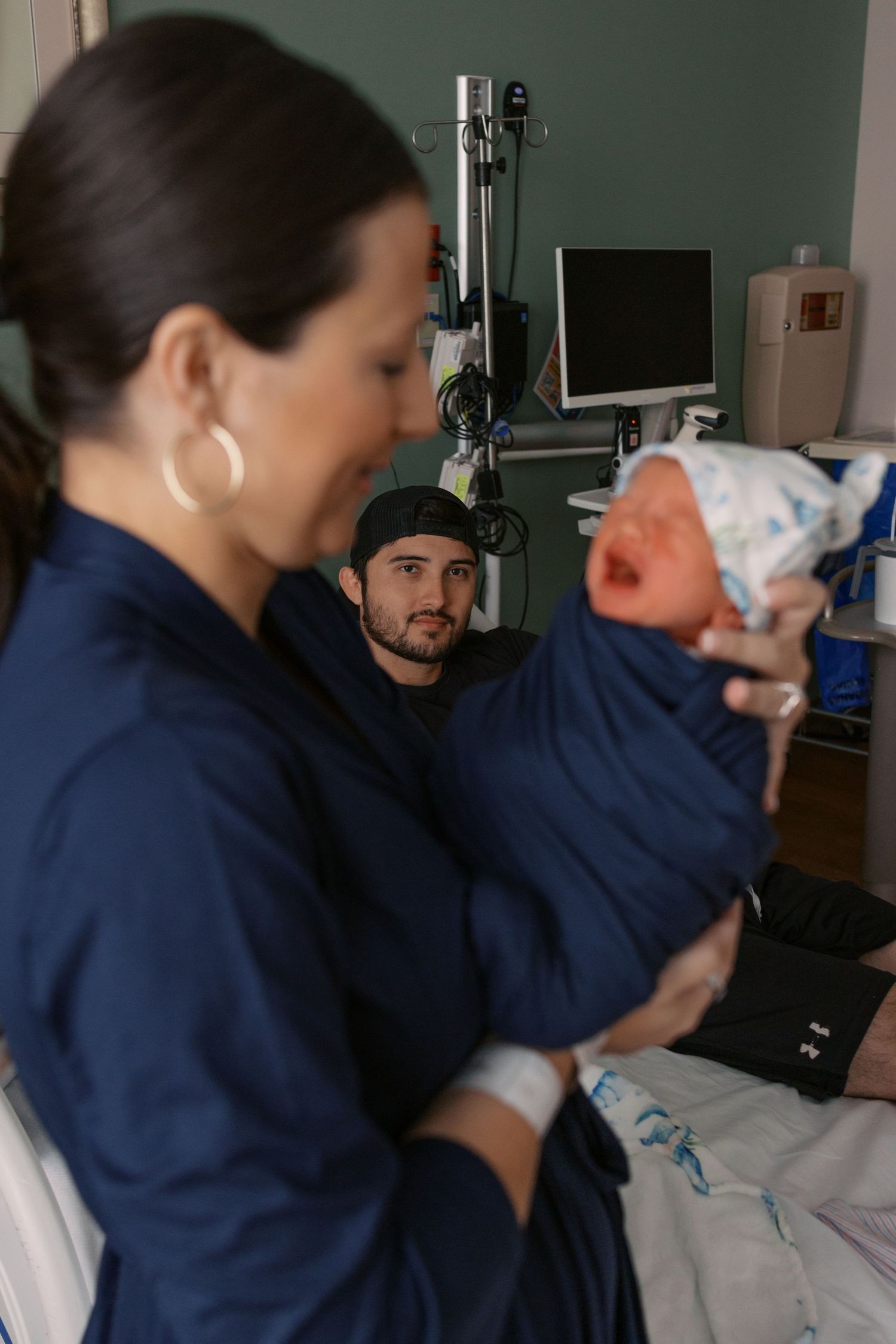 New parents in hospital room with mother holding crying newborn as father looks on.