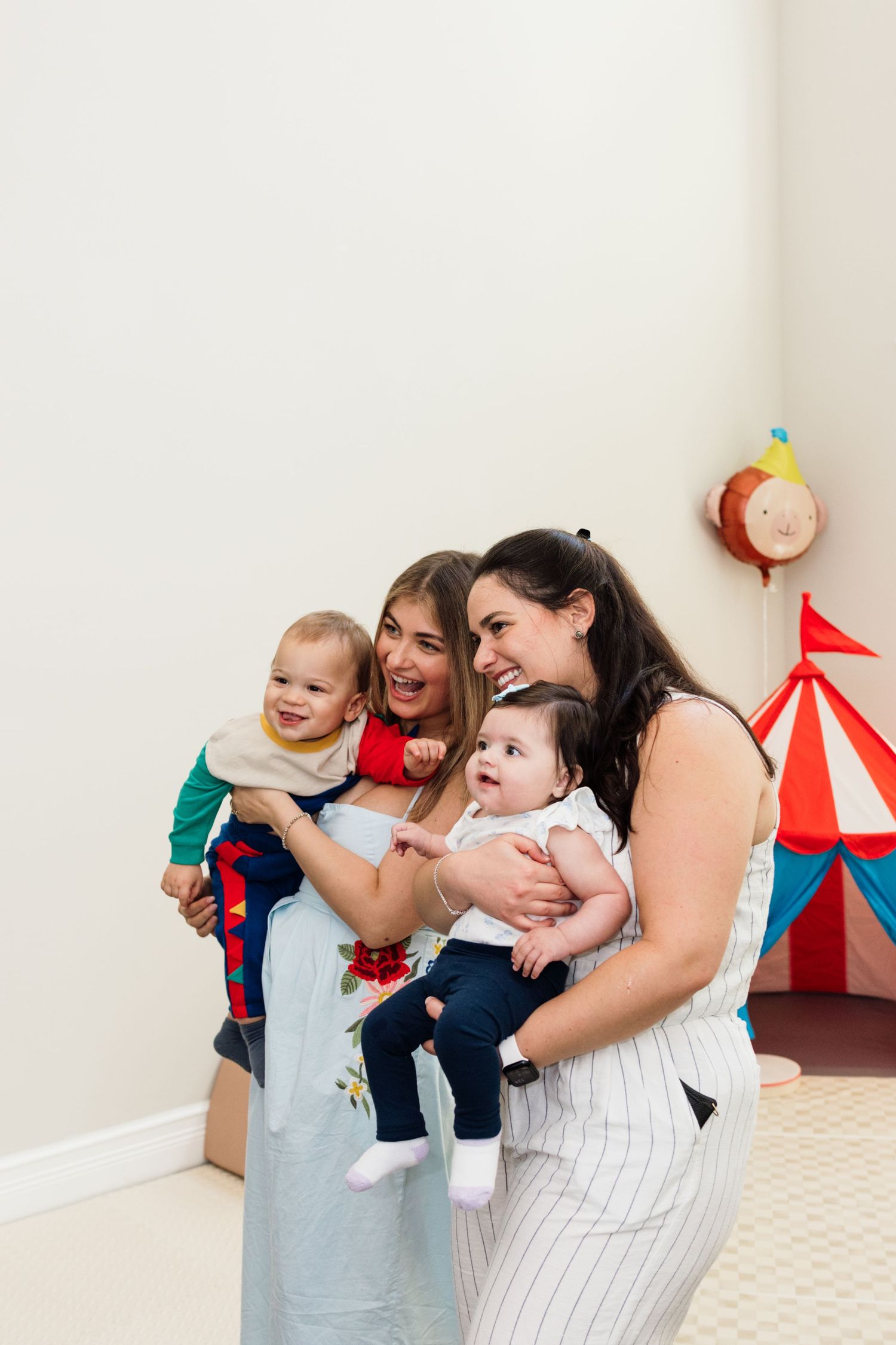 Two women smiling and holding babies at a circus-themed birthday party with colorful tent decorations.