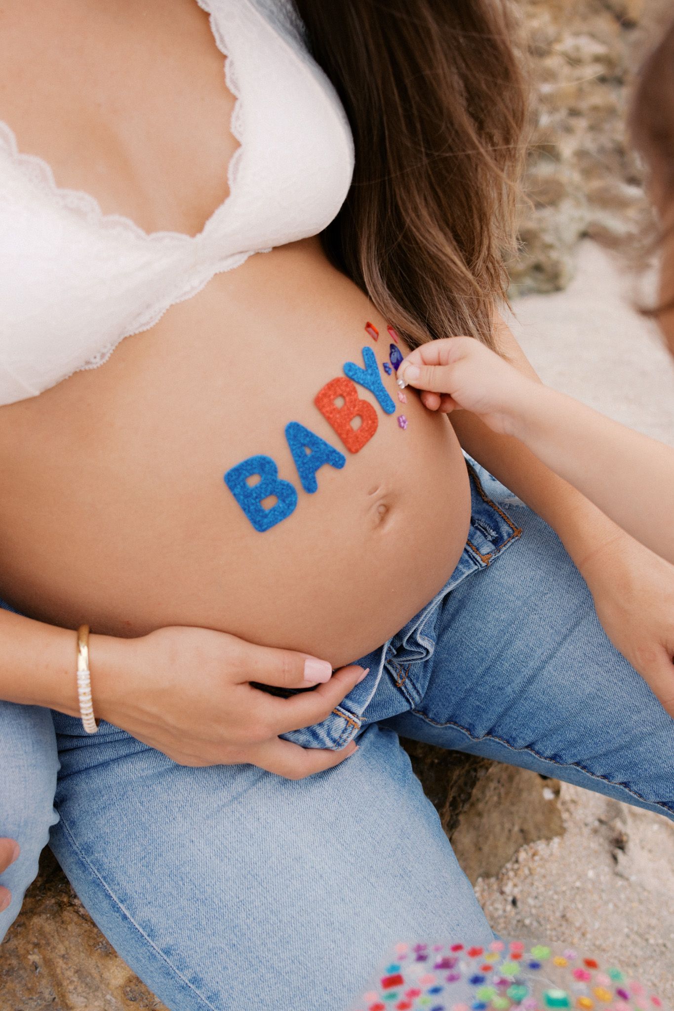 Pregnant woman with colorful BABY letters painted on her baby bump, wearing white bra and jeans.