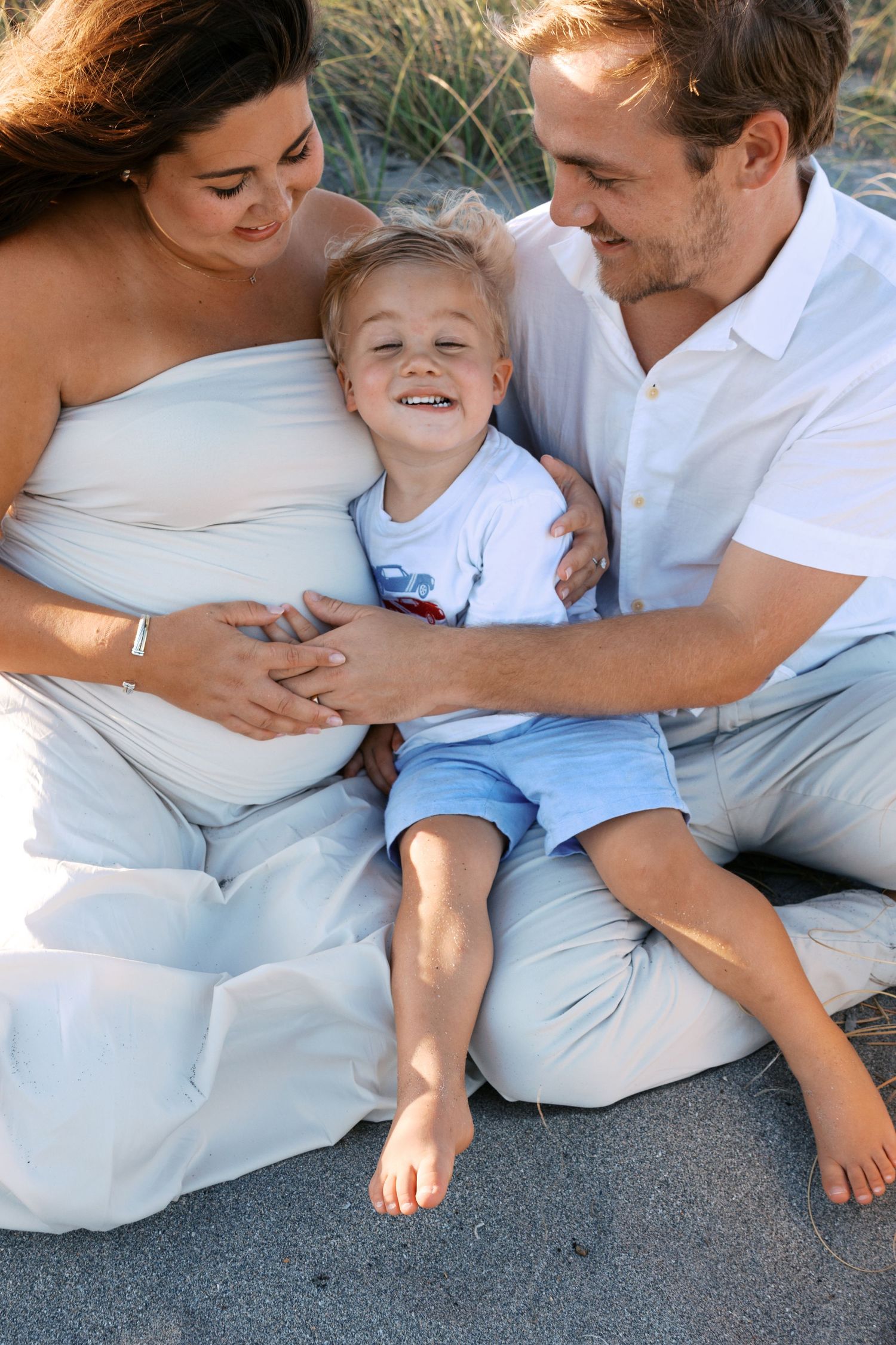 Happy toddler boy sits between pregnant mother and father on sandy beach during family maternity photo session.