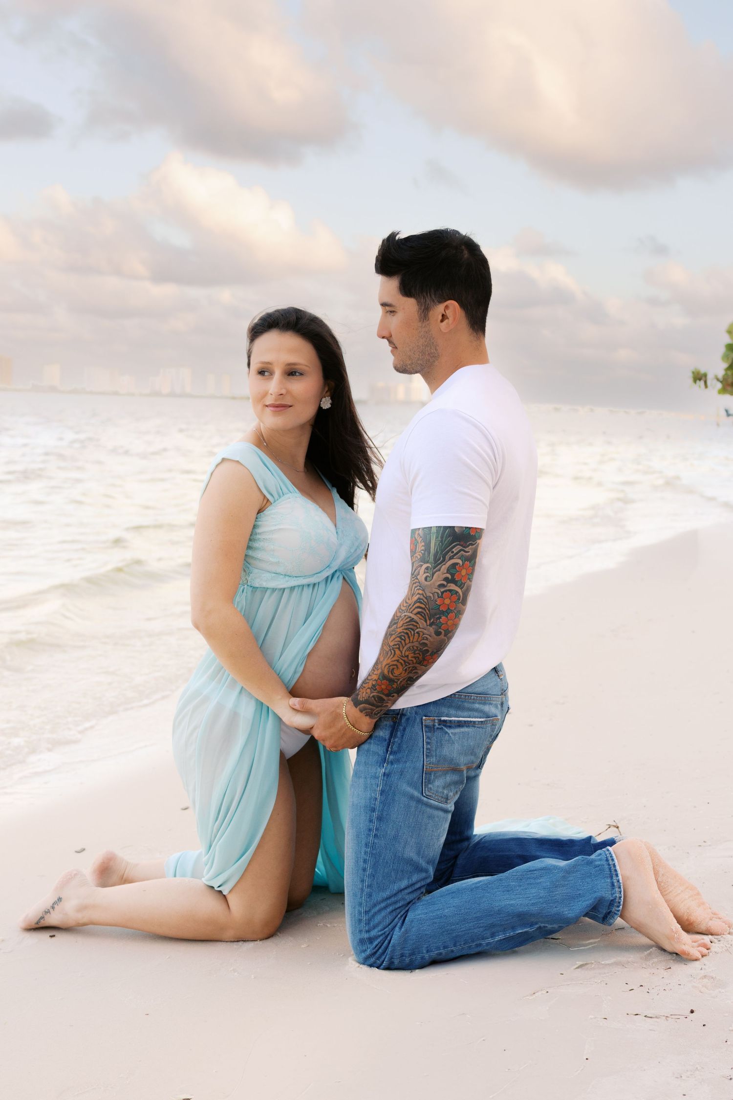 Pregnant couple posing on sandy beach, man kneeling in jeans with tattoo sleeve, woman in teal maternity dress.