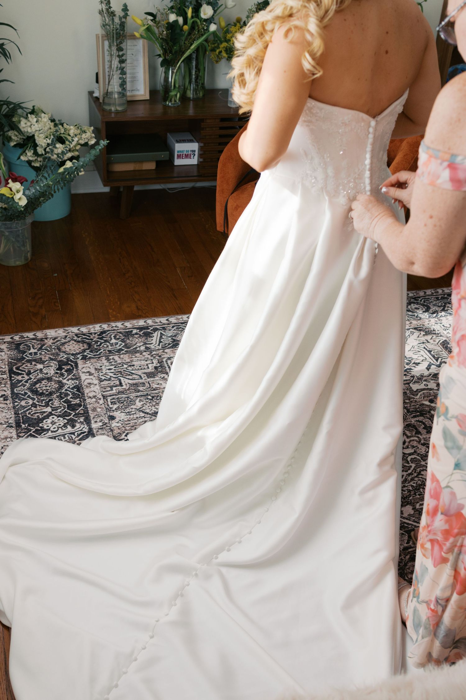 Bridesmaid fastening buttons on back of elegant white satin wedding gown with train on bride with blonde curly hair.