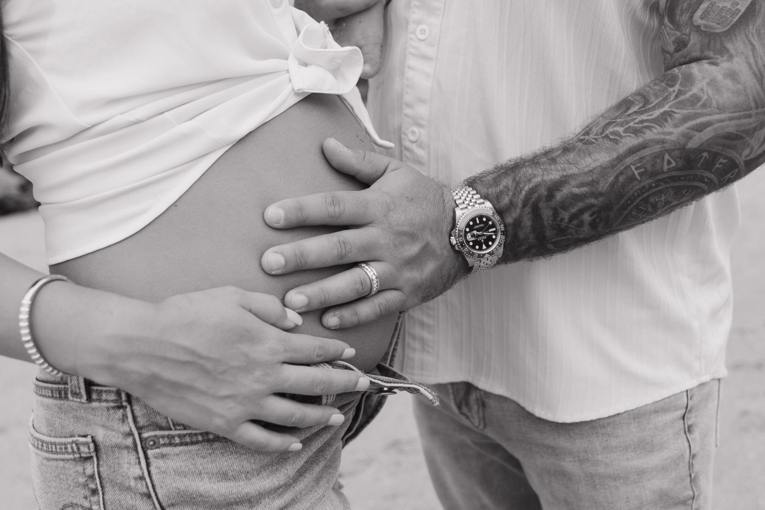 Black and white photo of a pregnant woman with her partner's tattooed hand resting on her baby bump.
