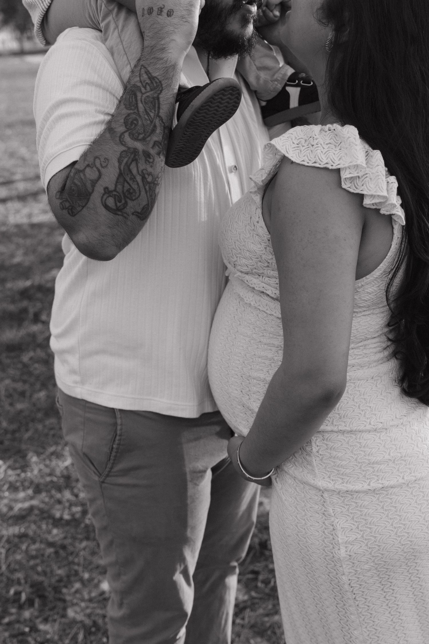 Black and white photo of a pregnant couple outdoors, man with tattoos holding a baby shoe near womans baby bump.