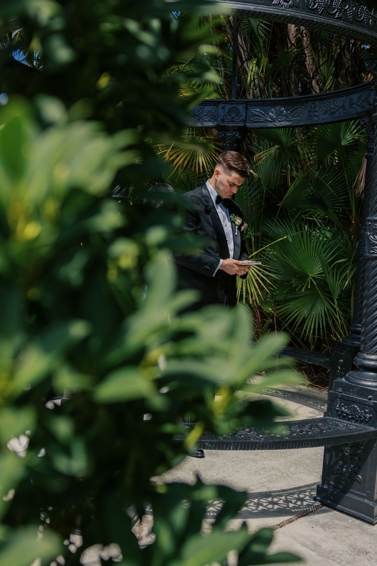Groom in tuxedo reading vows among lush tropical greenery and ornate black iron gazebo.