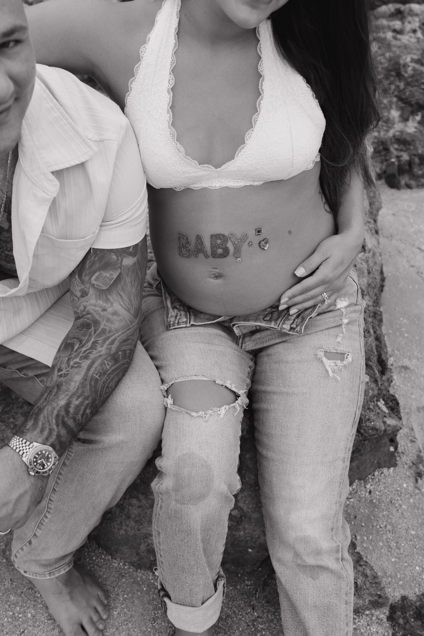 Black and white photo of pregnant womans baby bump with BABY letters, beside tattooed partner.