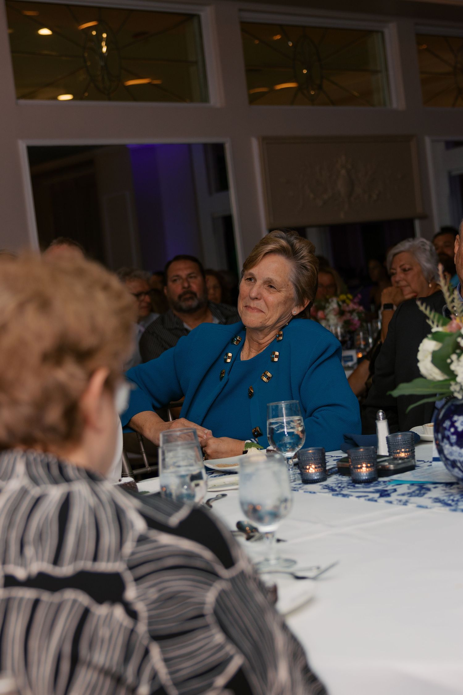 Elderly woman in teal jacket seated at elegant banquet table with wine glasses and candles at formal dinner event.