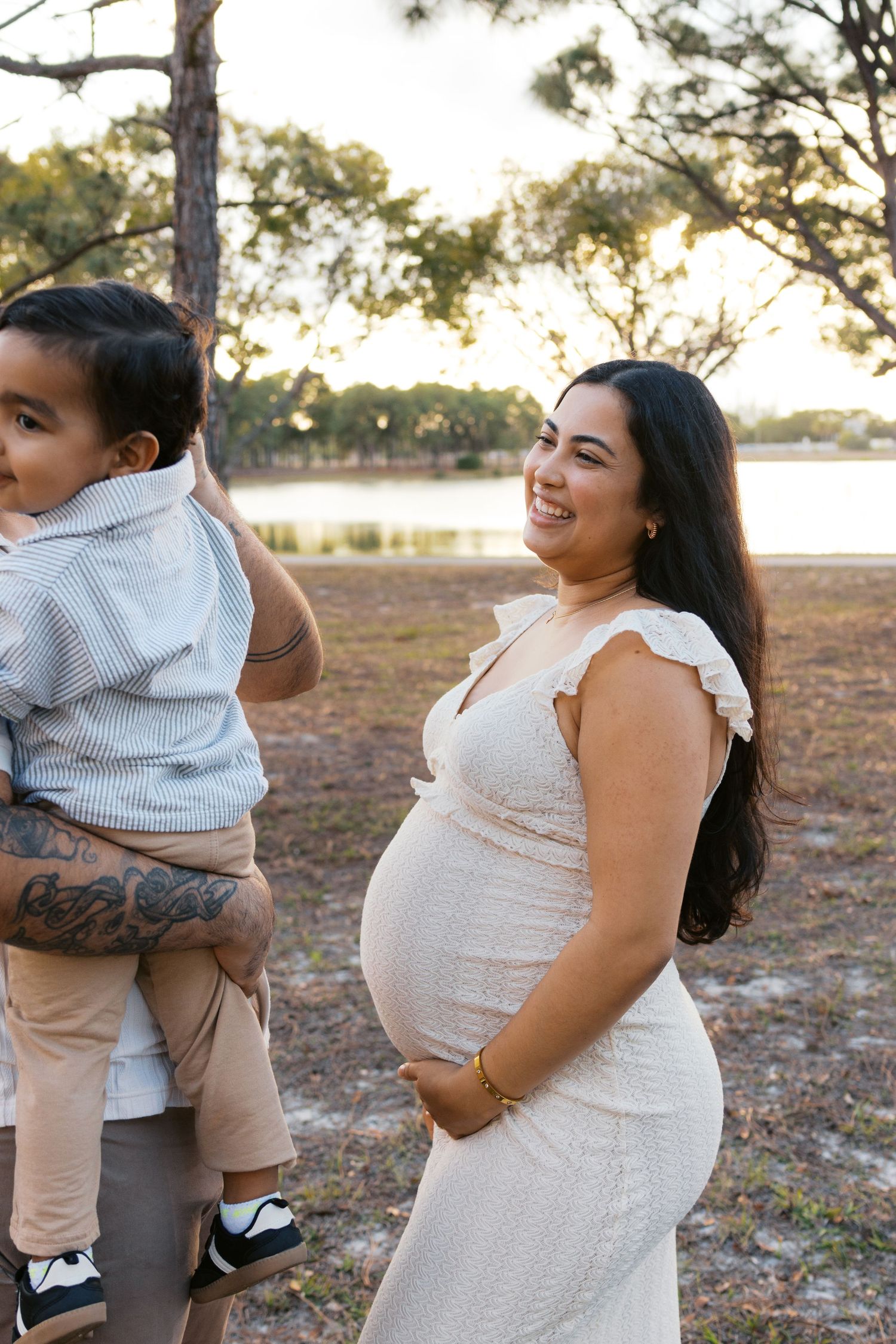 Smiling pregnant woman in white dress holding her baby bump outdoors near a lake at golden hour.