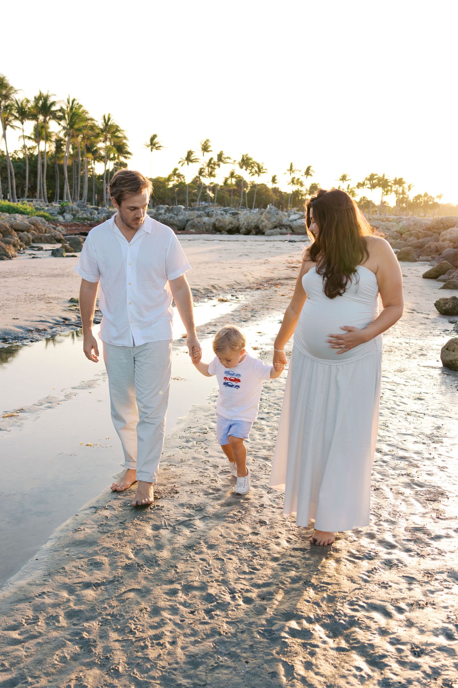 Pregnant woman in white dress walks on tropical beach at sunset with husband and young toddler child.