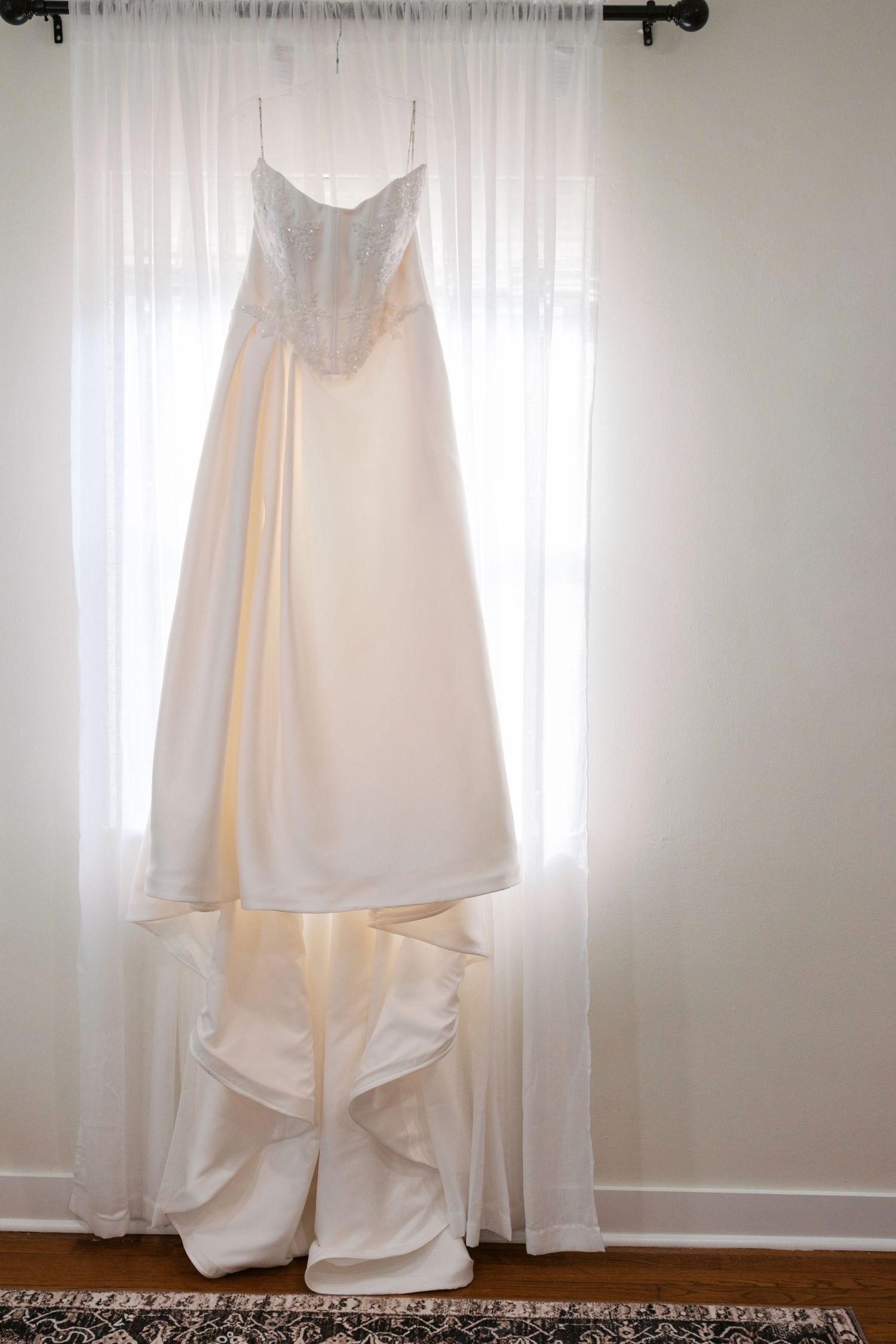 Elegant white strapless wedding gown with lace bodice hanging by window in natural light.