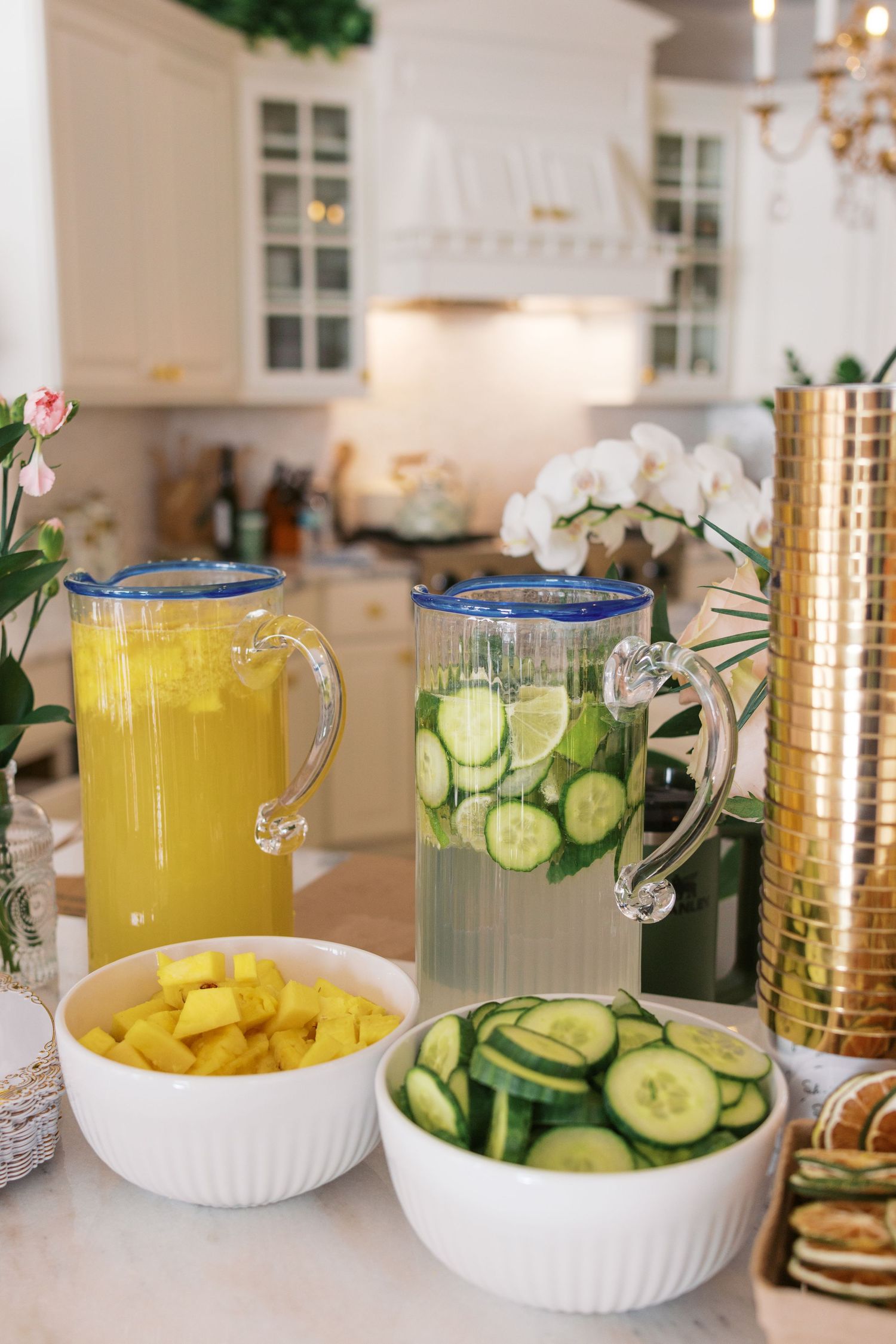 Two glass pitchers with blue rims filled with lemonade and cucumber water, beside bowls of mango and cucumber slices.