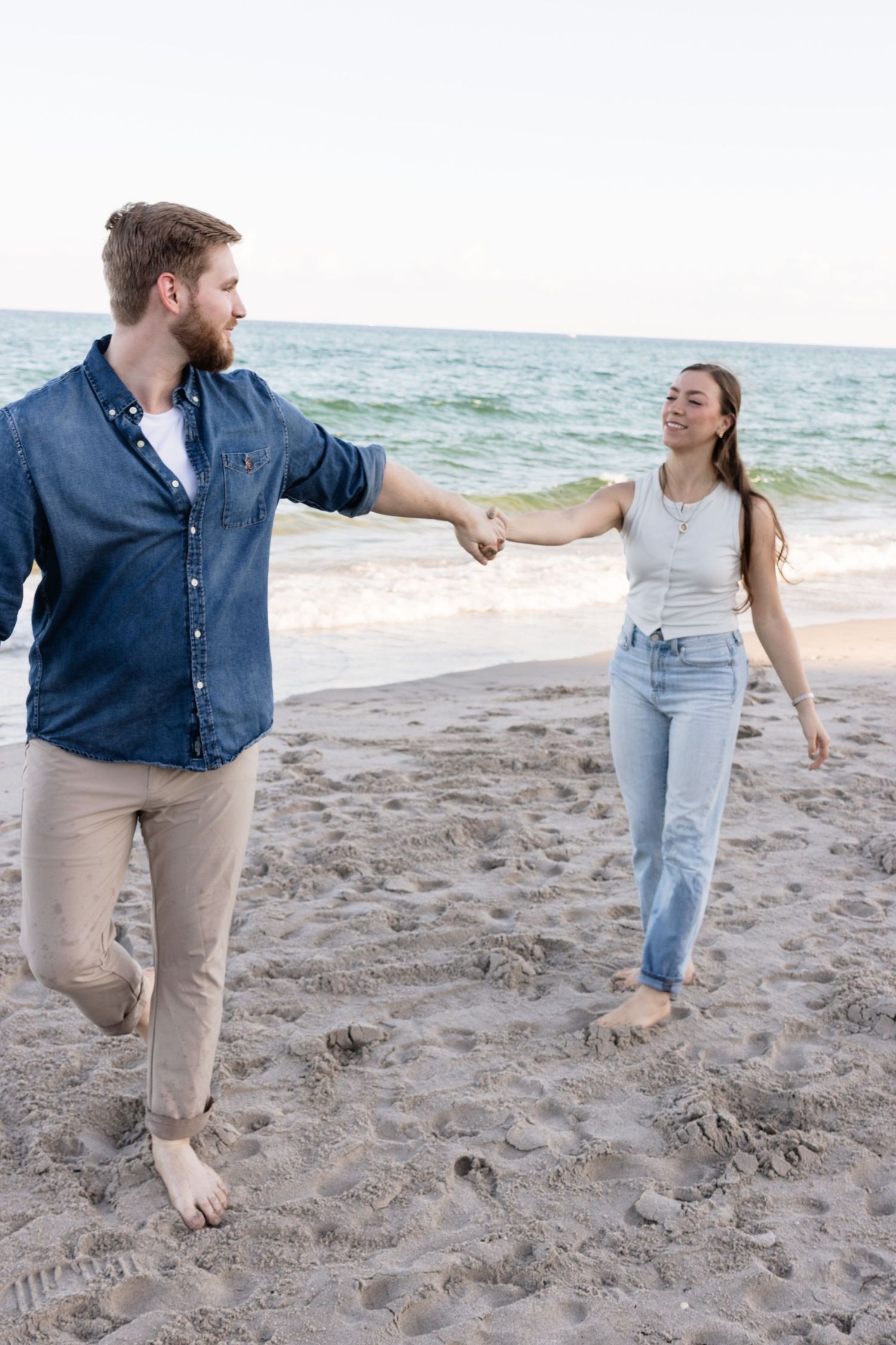 A couple playfully walking and holding hands on a sandy beach at sunset.