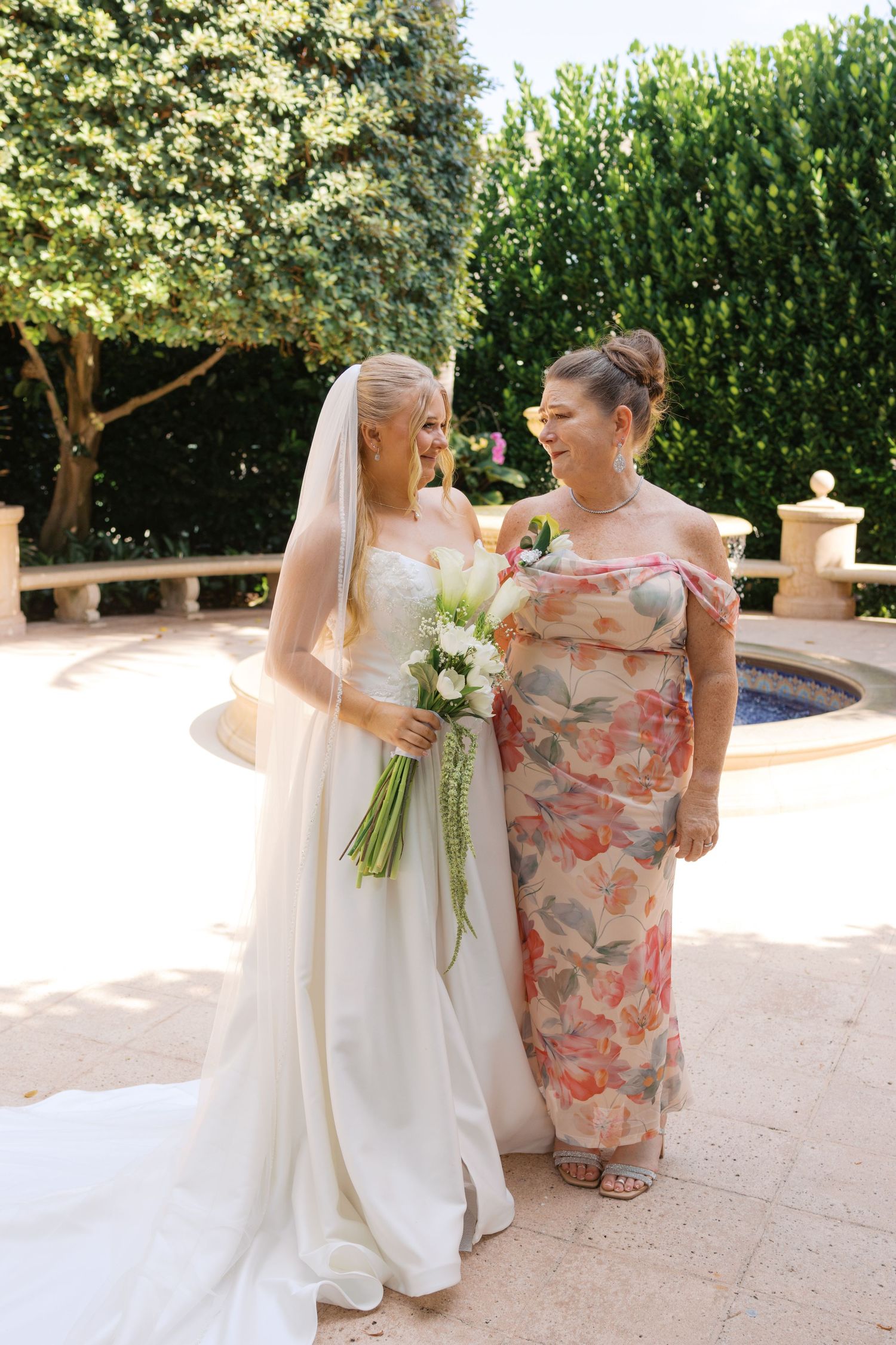 Blonde bride in strapless white gown holding floral bouquet stands with woman in floral dress at outdoor wedding.