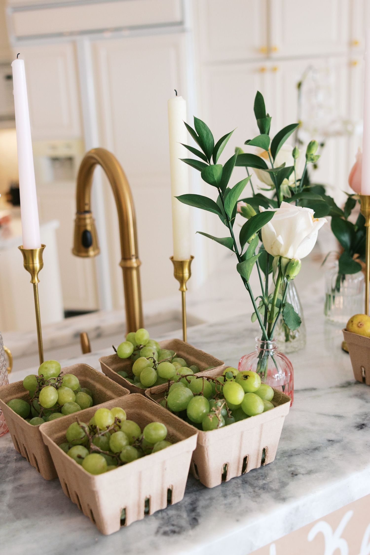 Green grapes in cardboard berry baskets on marble counter with white roses, candles, and gold faucet in elegant kitchen.