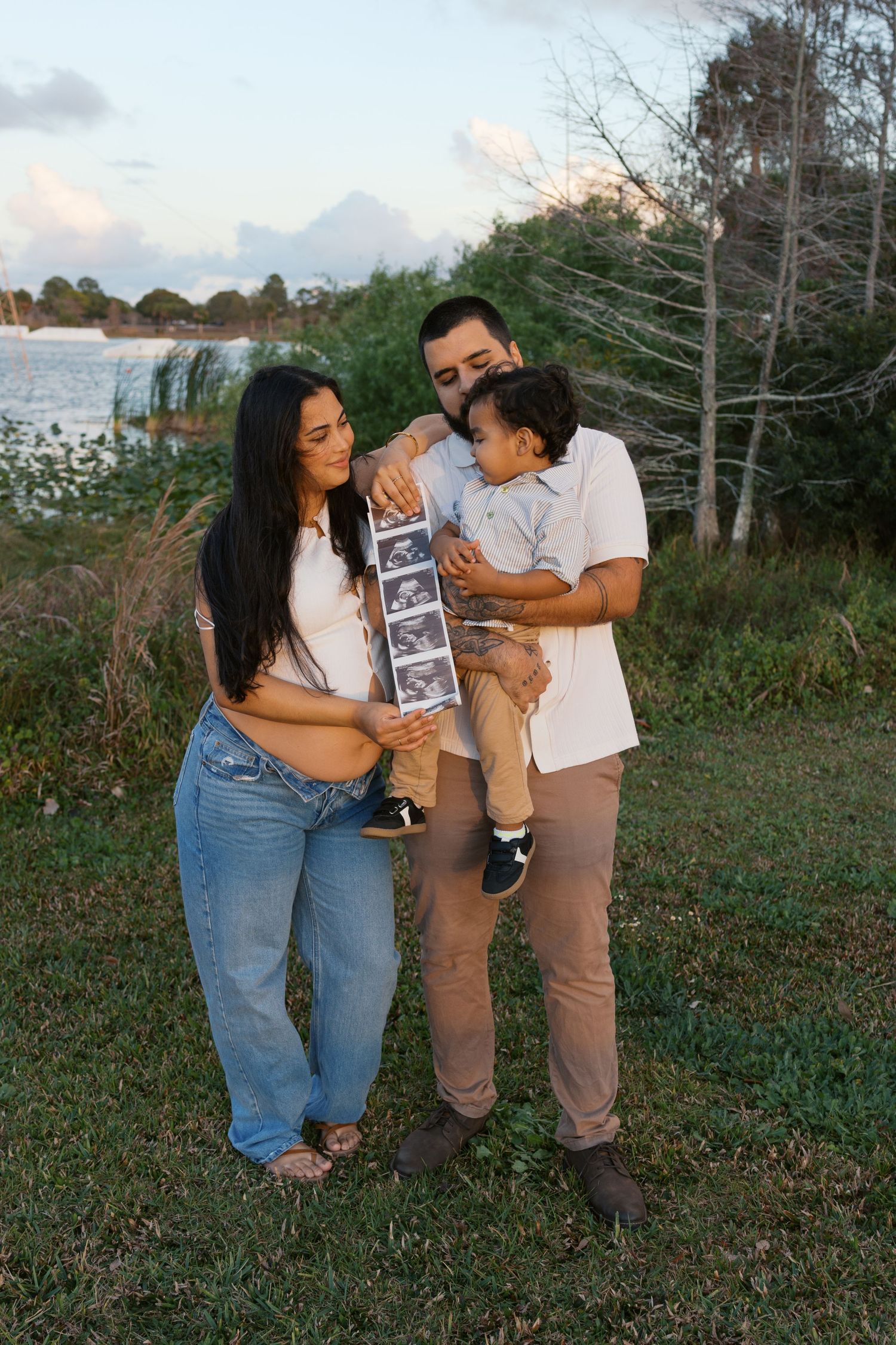 Pregnant woman and man holding toddler examine ultrasound photos outdoors near a lake at sunset.