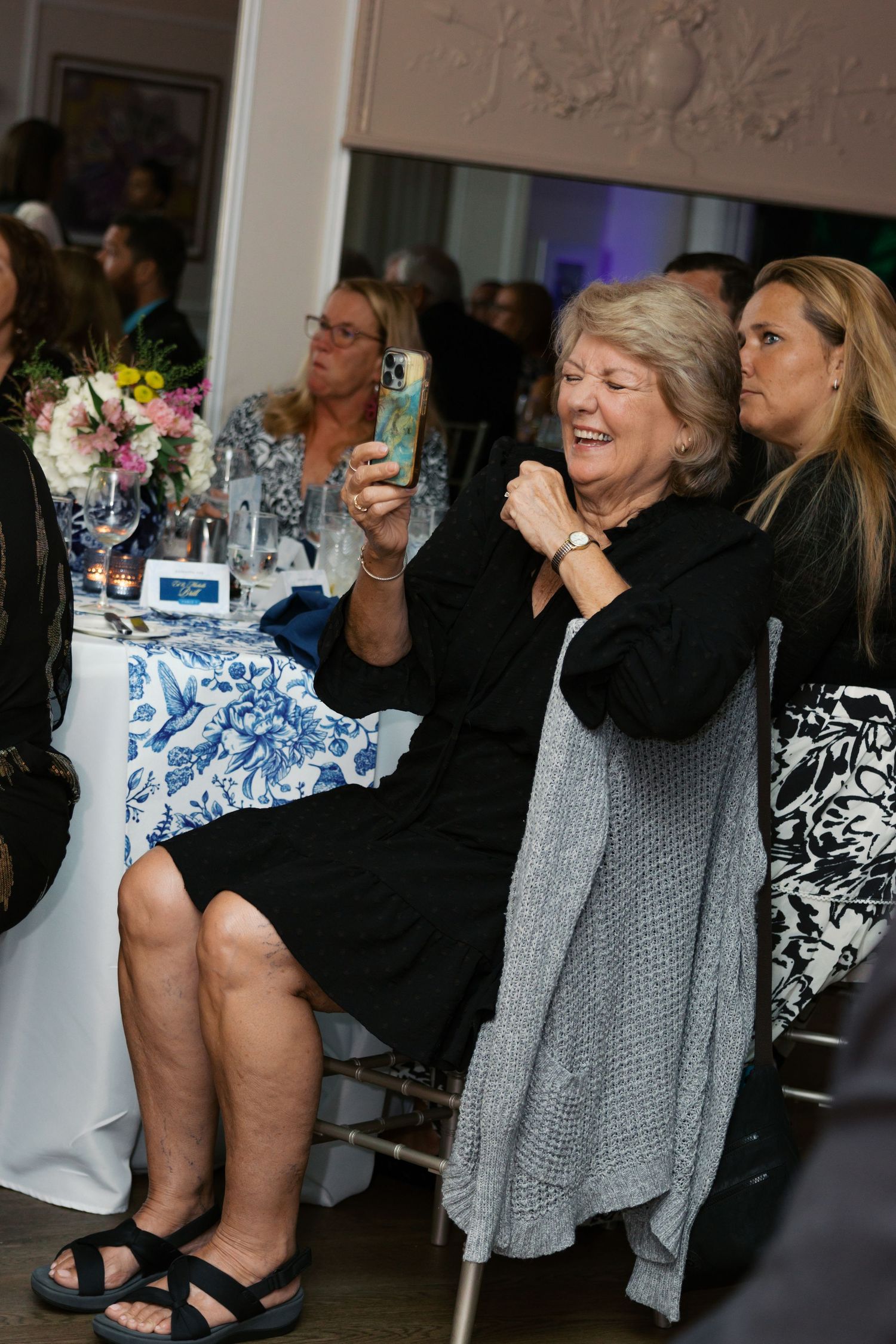 A woman in black outfit laughs while taking a photo on her phone at an elegant indoor event.