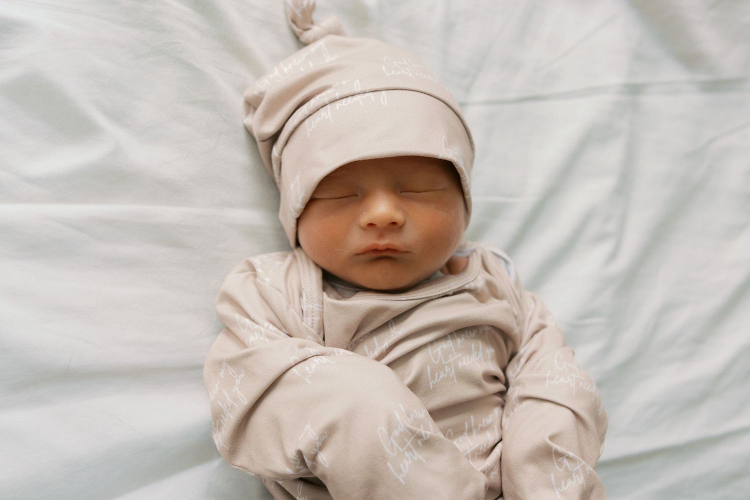 Newborn baby sleeping peacefully in beige outfit and matching knot hat on white bedding.