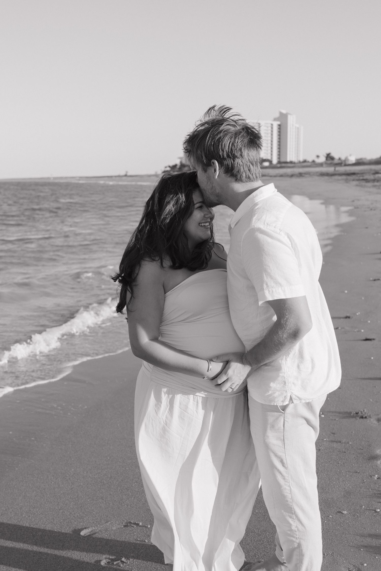 Black and white photo of a romantic couple embracing and smiling on a sandy beach with ocean waves and a hotel in the background.