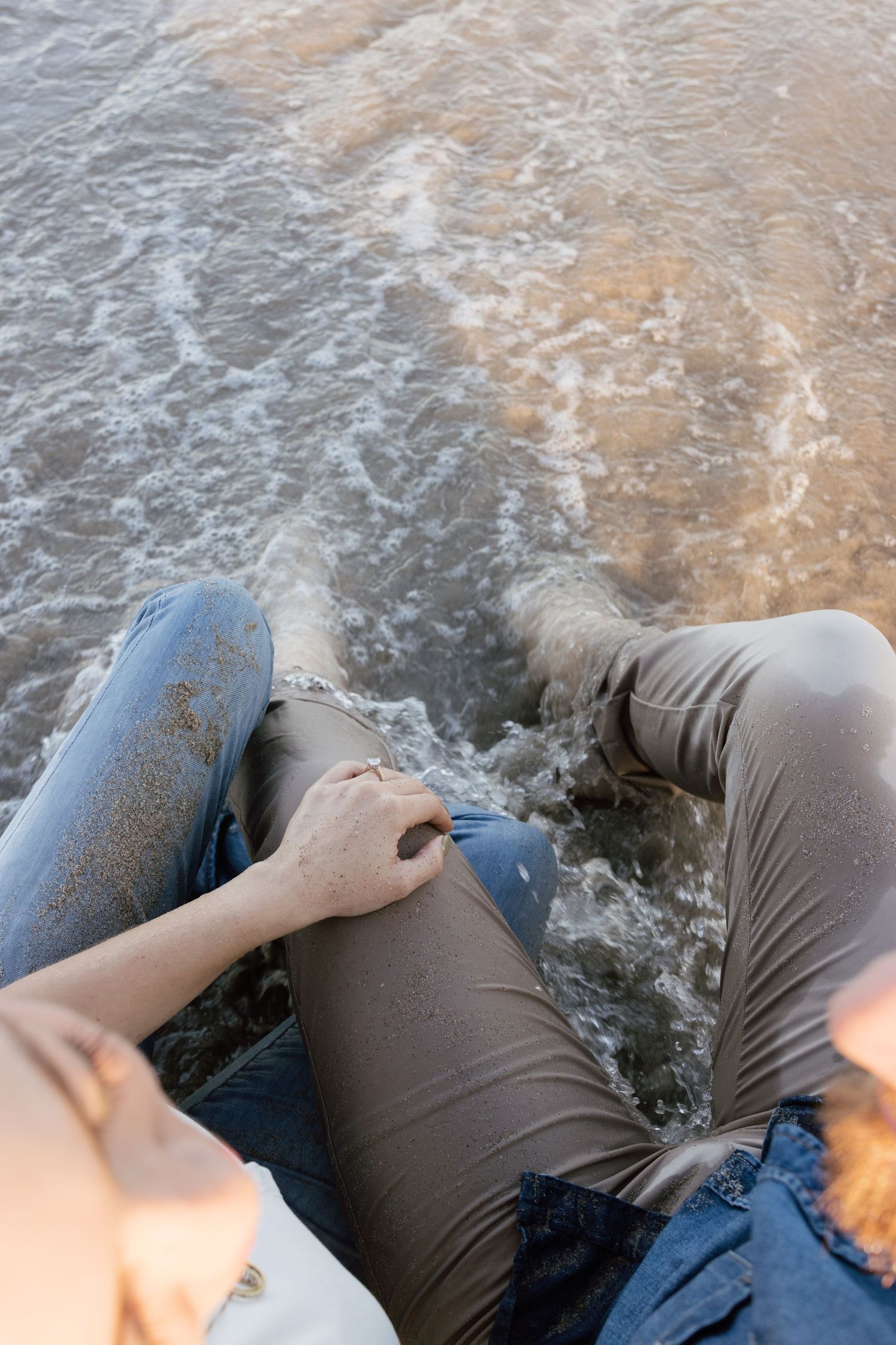 Waves washing over couple lying on beach in casual clothing.