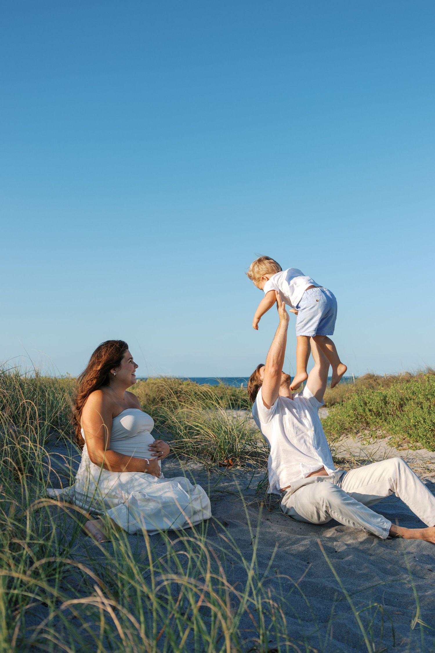 Pregnant woman sits on beach dunes while partner lifts toddler in air, family dressed in white near ocean.