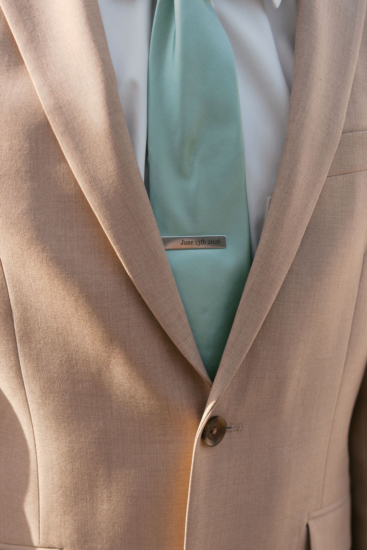 Beige wedding suit with mint green tie and personalized silver tie clip in sunlight.