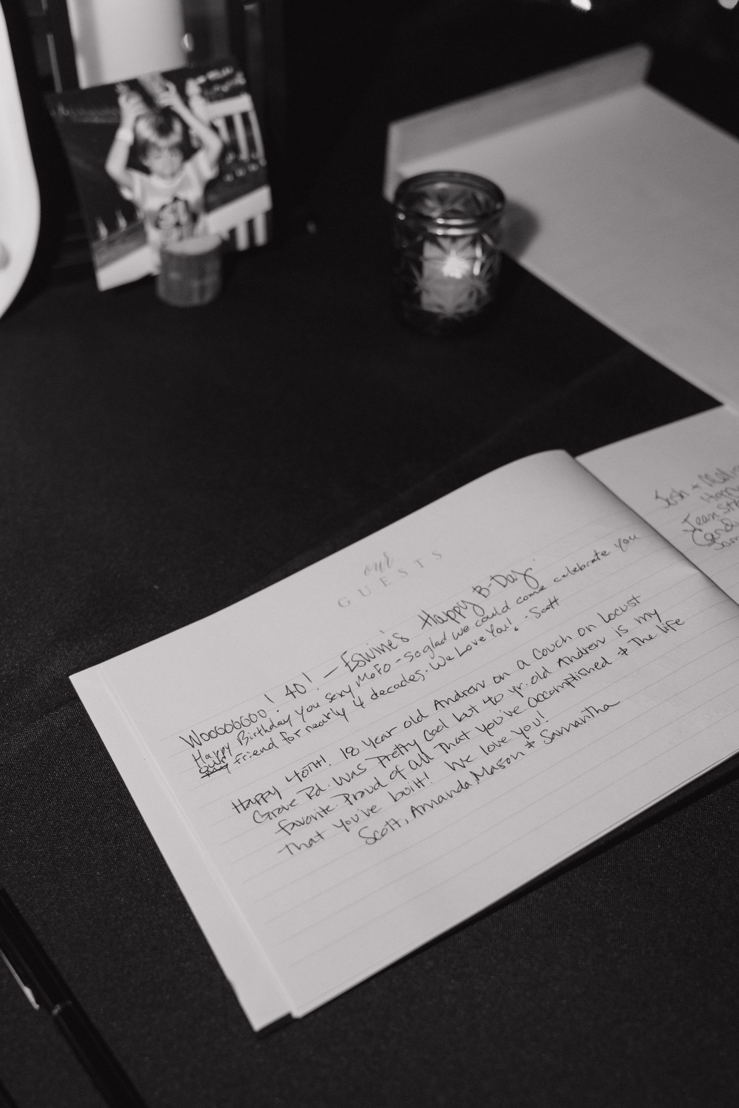 Black and white photo of an open guest book with handwritten birthday messages on a dark table with a candle.