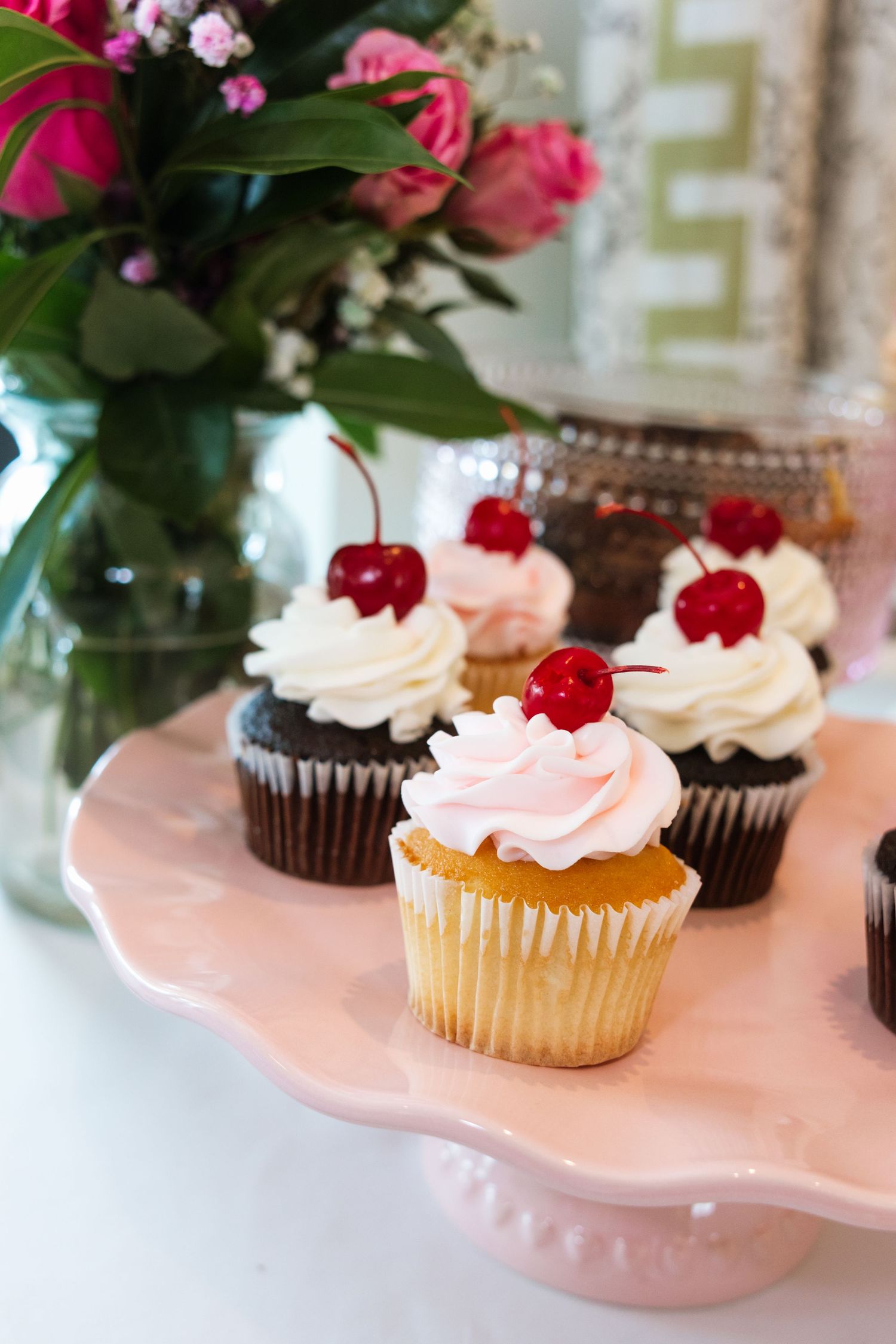 Assorted cupcakes with white and pink frosting topped with red cherries displayed on a pink cake stand.