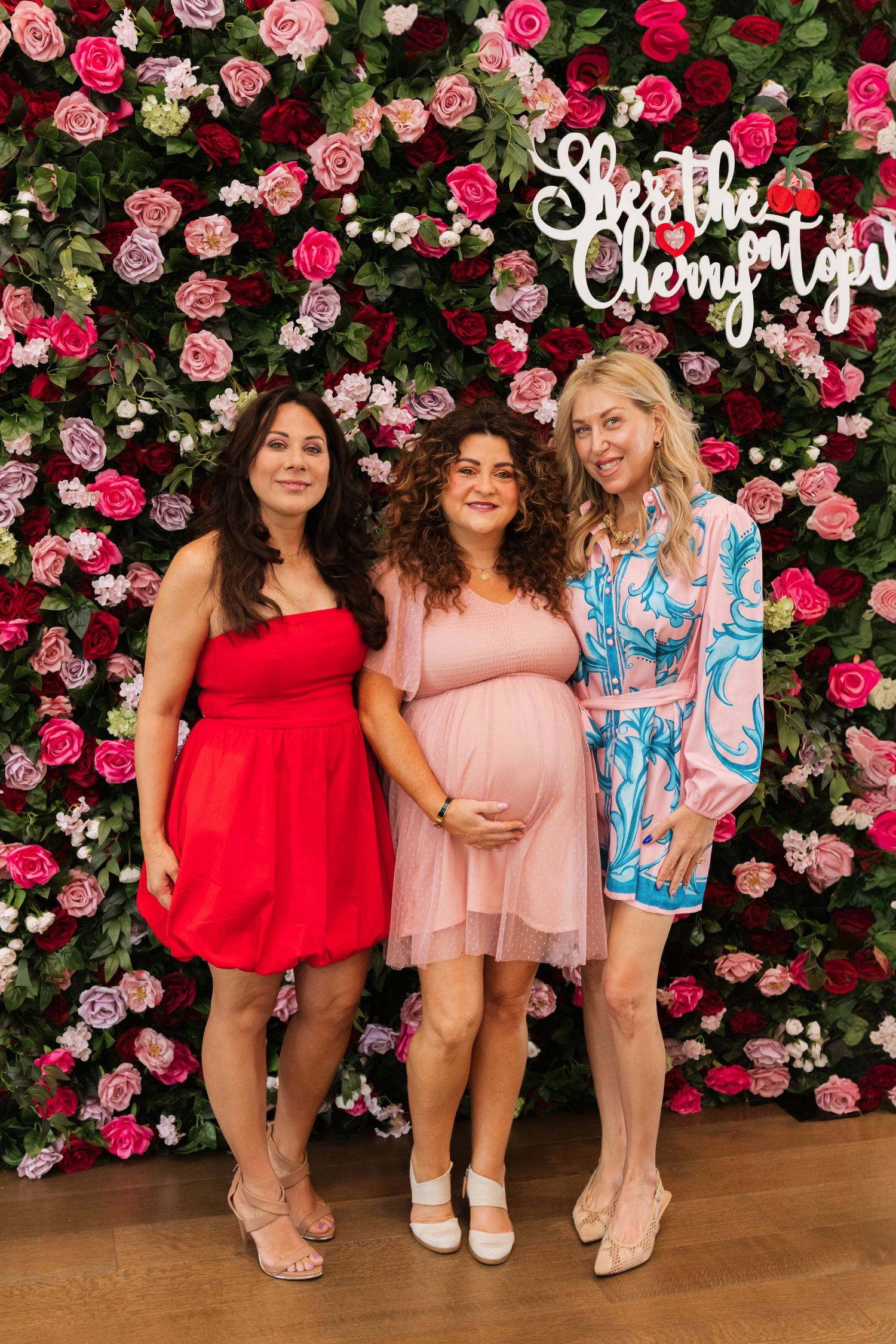 Three women posing in front of a floral wall backdrop at a baby shower, with the pregnant guest of honor in pink.