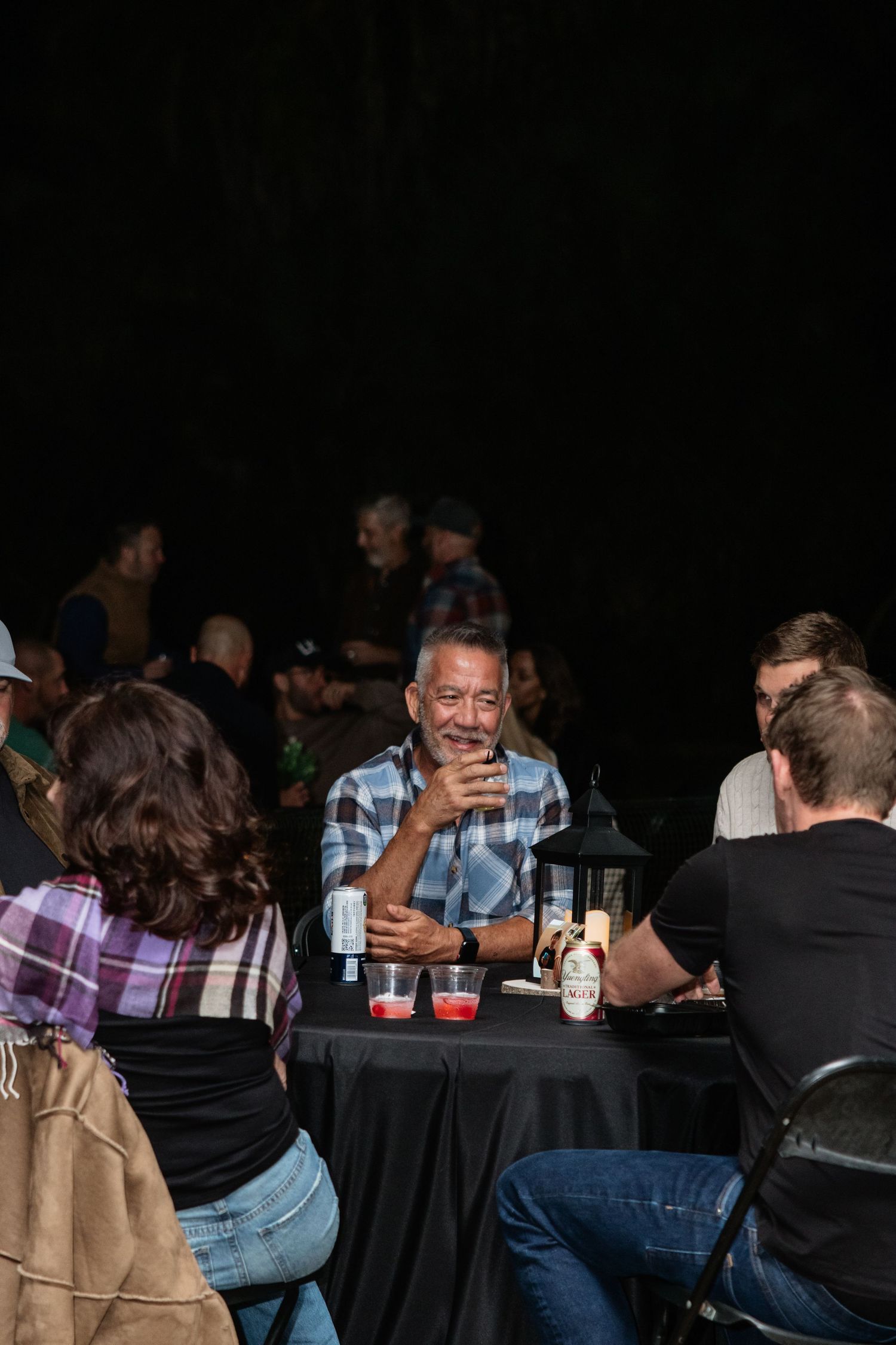 Group of adults socializing at a dark outdoor evening event, smiling man in plaid shirt holding a drink at a table.