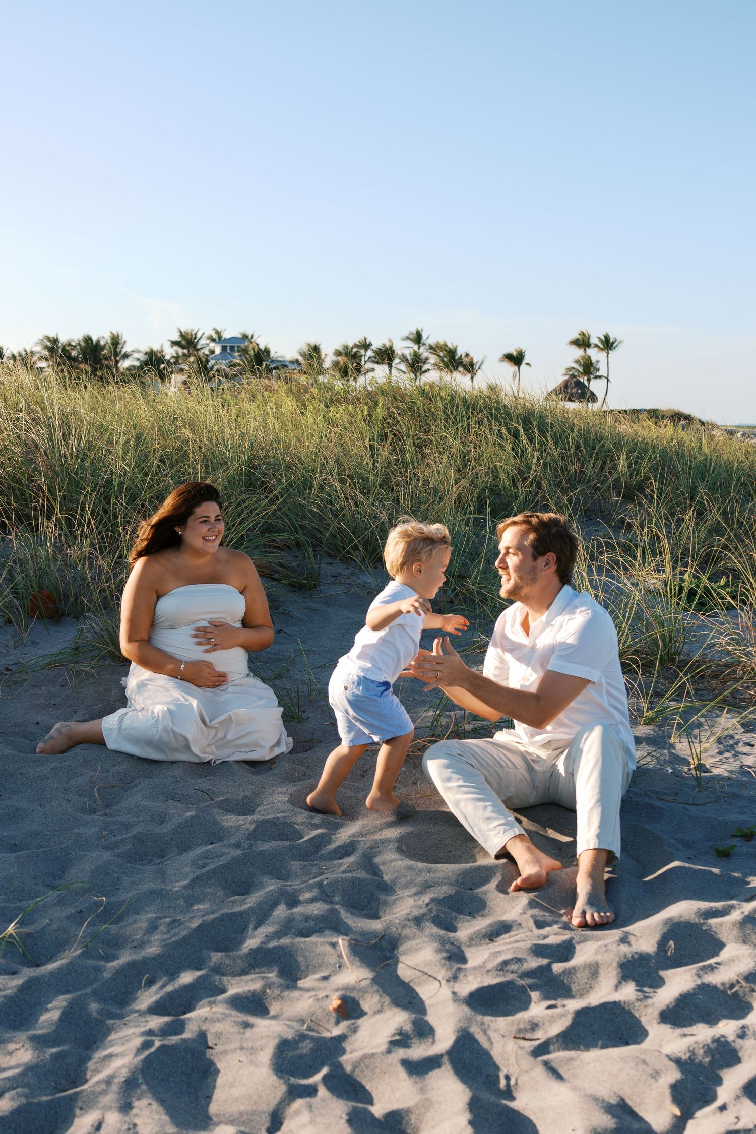 Joyful family sitting on sandy beach dunes, pregnant mother in white dress watching father play with young blond son.
