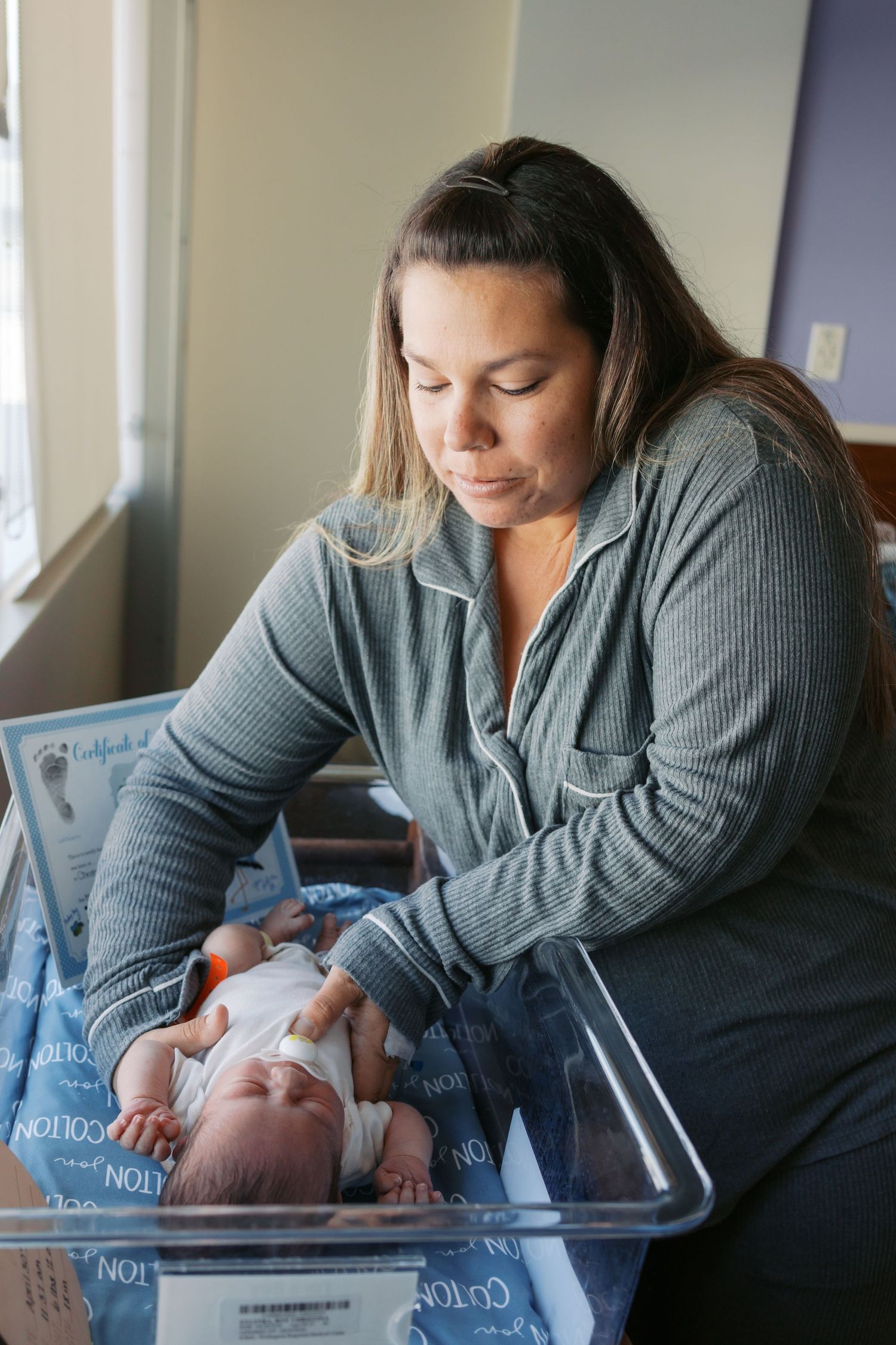 A mother in gray pajamas lovingly gazes at her newborn baby lying in a hospital bassinet.