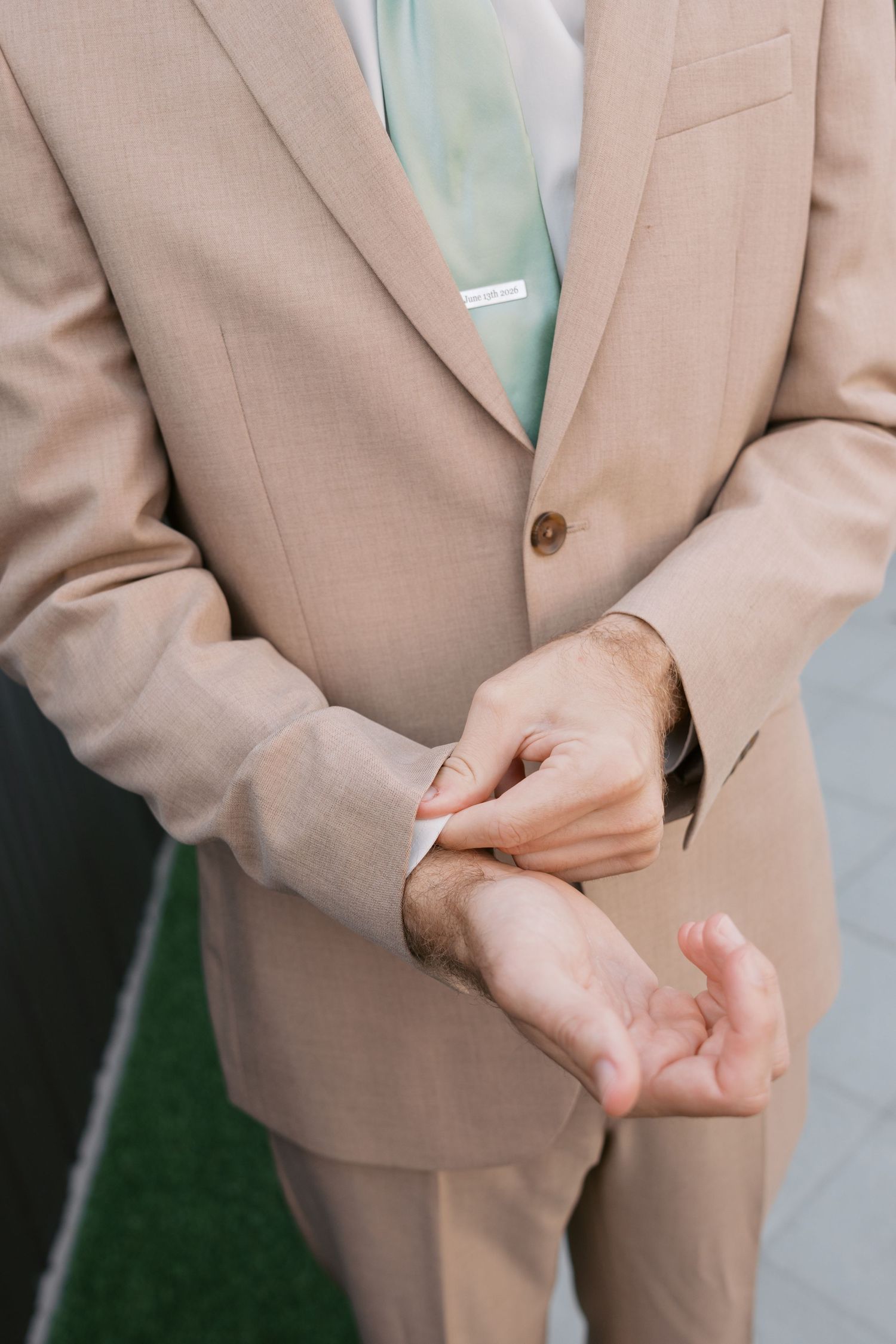 Groom in beige suit adjusting his cufflinks, wearing a mint green tie at a wedding ceremony.