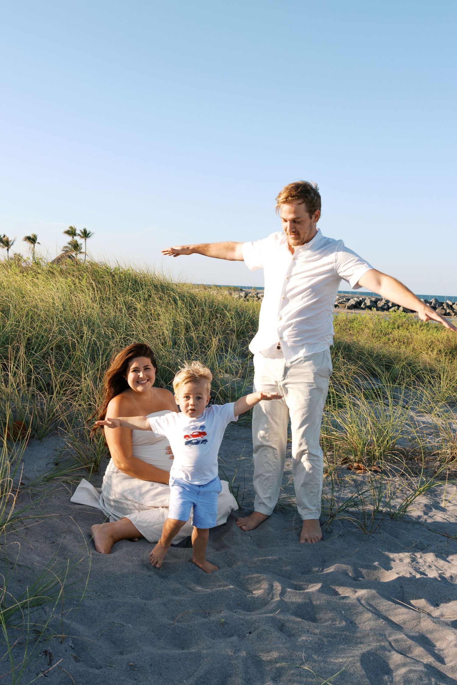 Family of three playing on sandy beach dunes, pregnant mother in white dress, father and toddler son enjoying sunny day.