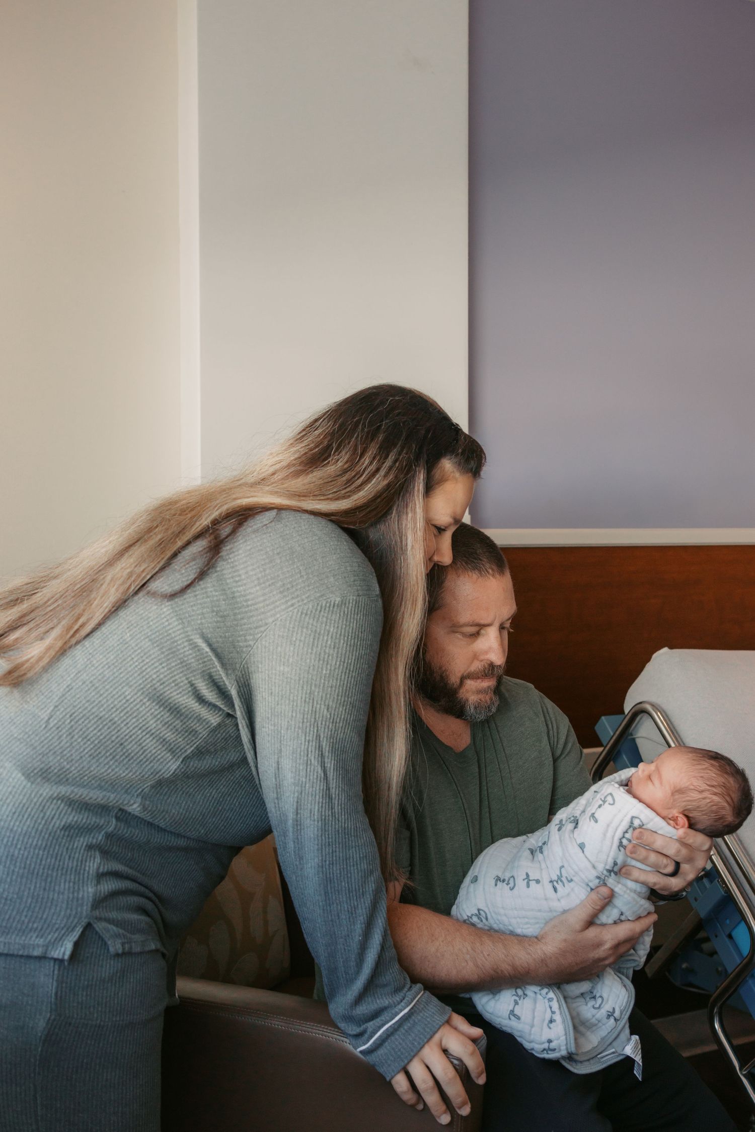 New parents tenderly gazing at their swaddled newborn baby in a hospital room setting.