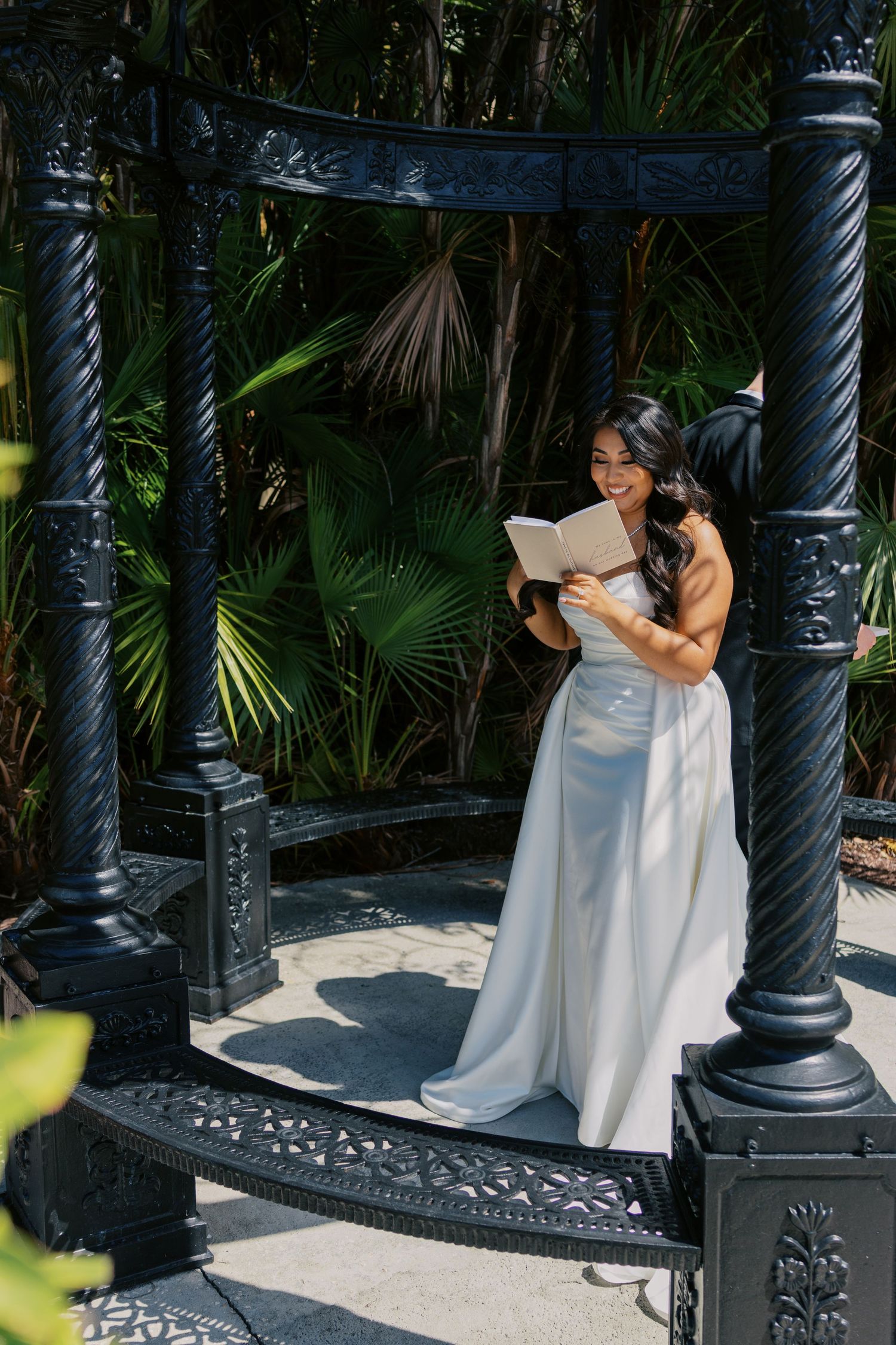 Bride in white gown reads vows under ornate black gazebo surrounded by tropical palms on sunny wedding day.