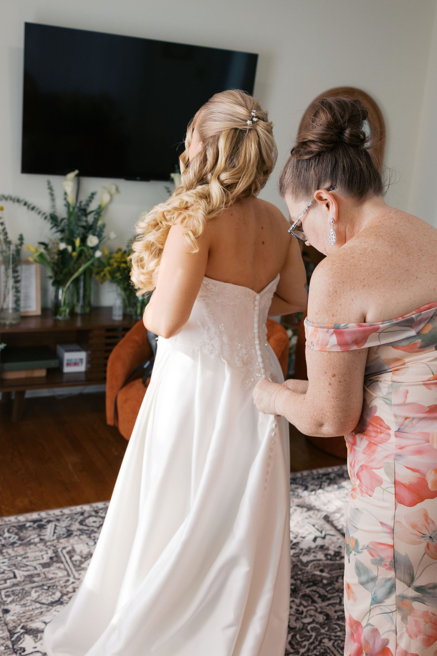 Bride in strapless white wedding gown being helped with buttons by woman in floral dress indoors.