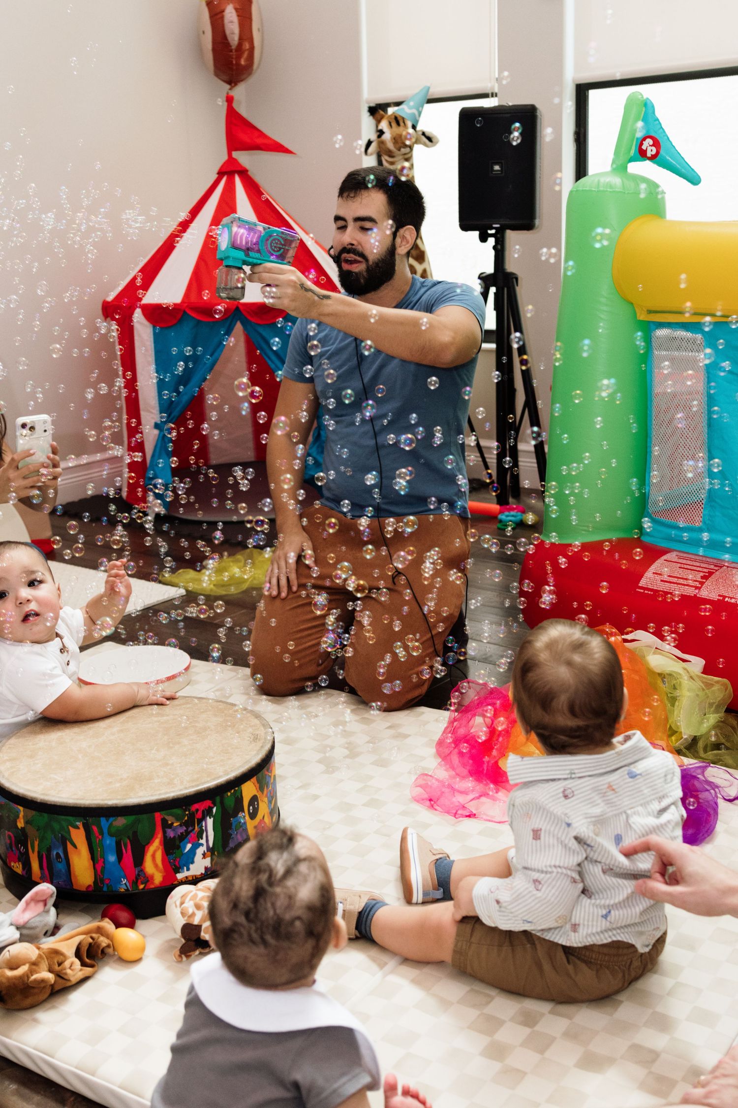 Man uses bubble gun to entertain babies at colorful circus-themed party with bouncy castle and drum.