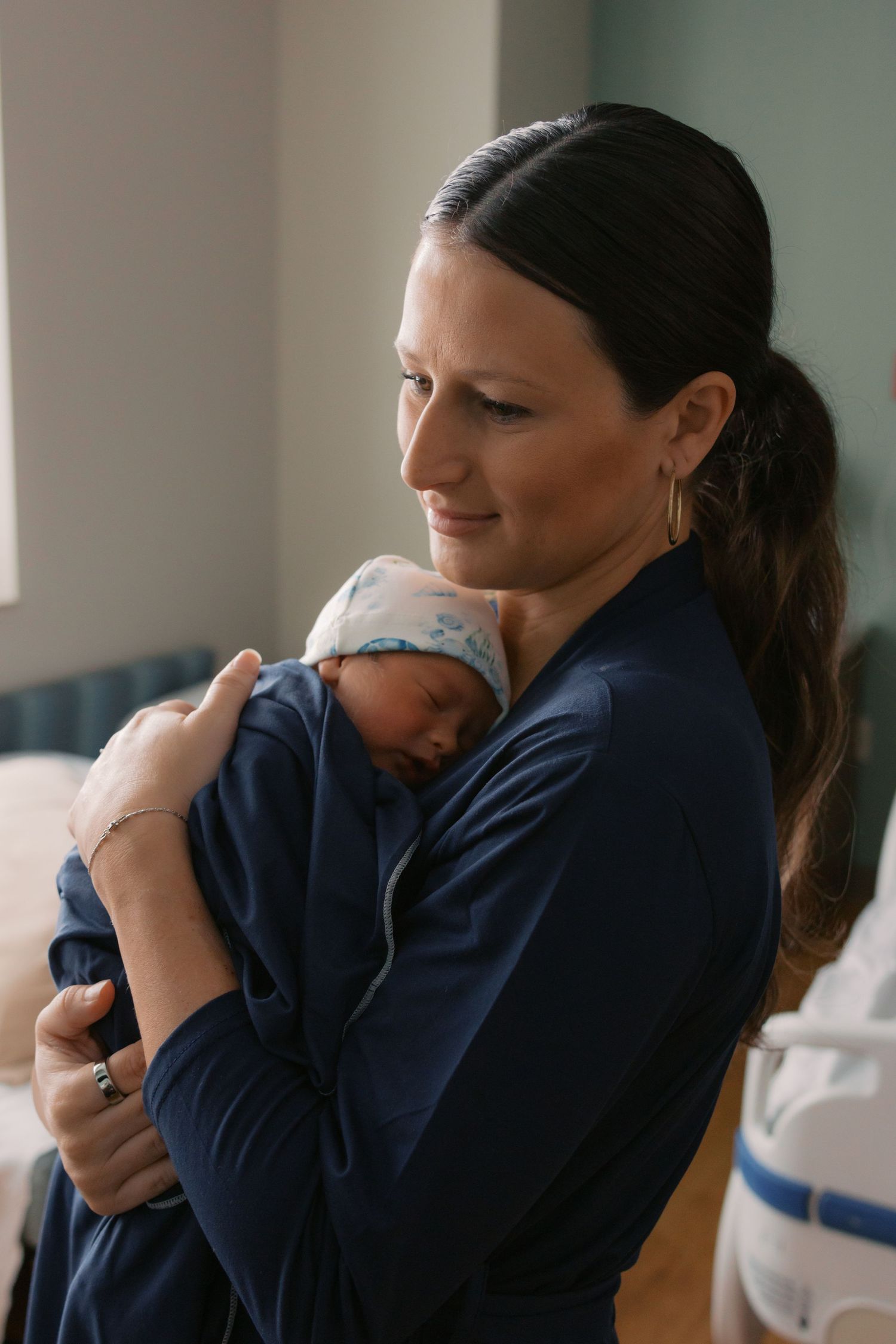 New mother in navy robe gently cradling her sleeping newborn baby against her chest in hospital room.