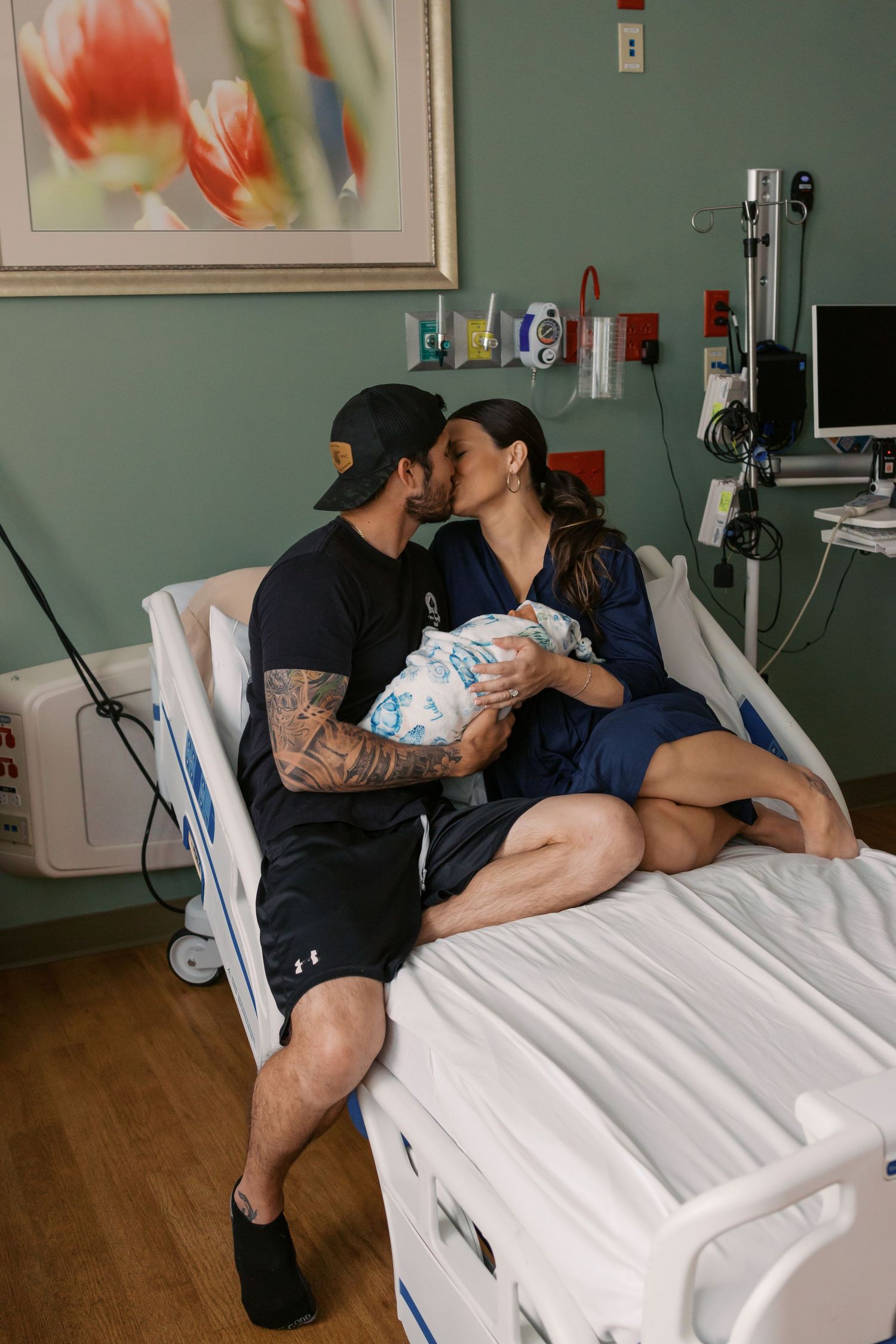 New parents share a tender kiss in hospital room while holding their newborn baby wrapped in blanket.