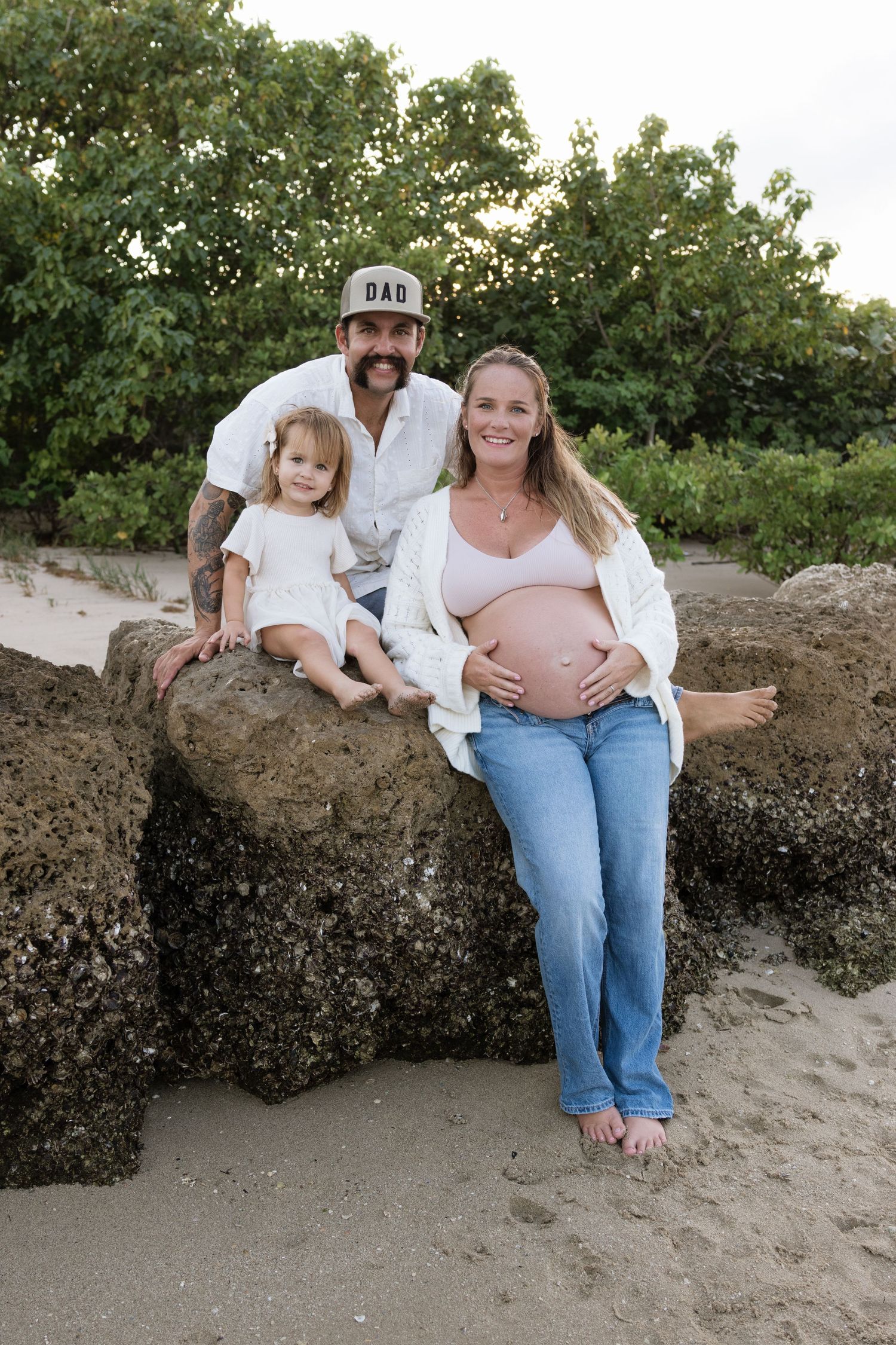 Pregnant woman poses with man in DAD cap and young girl on beach rocks with lush green trees behind them.