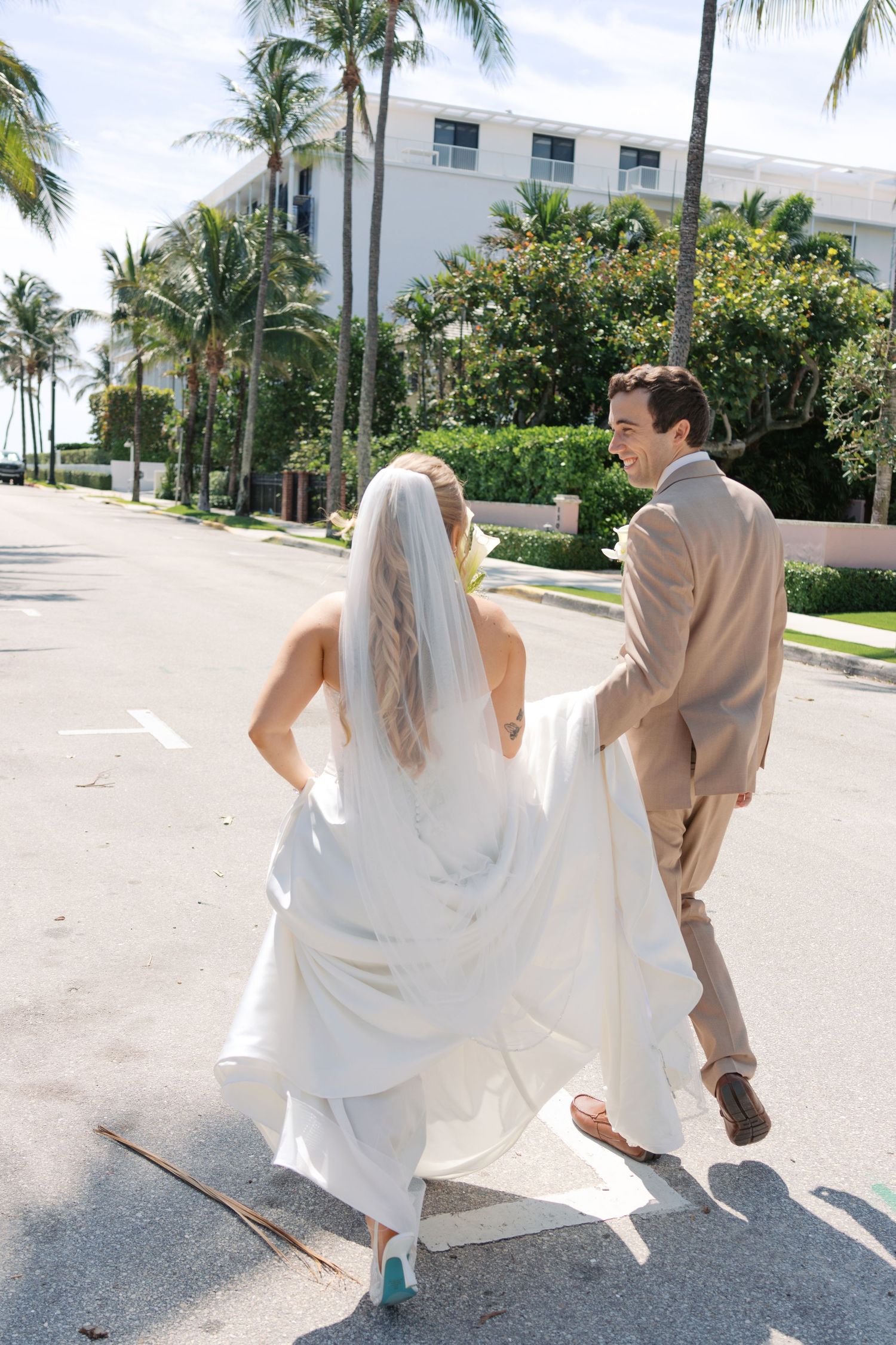 A bride and groom cross a sunny street lined with palm trees, the groom smiling while holding the bride's white gown train.