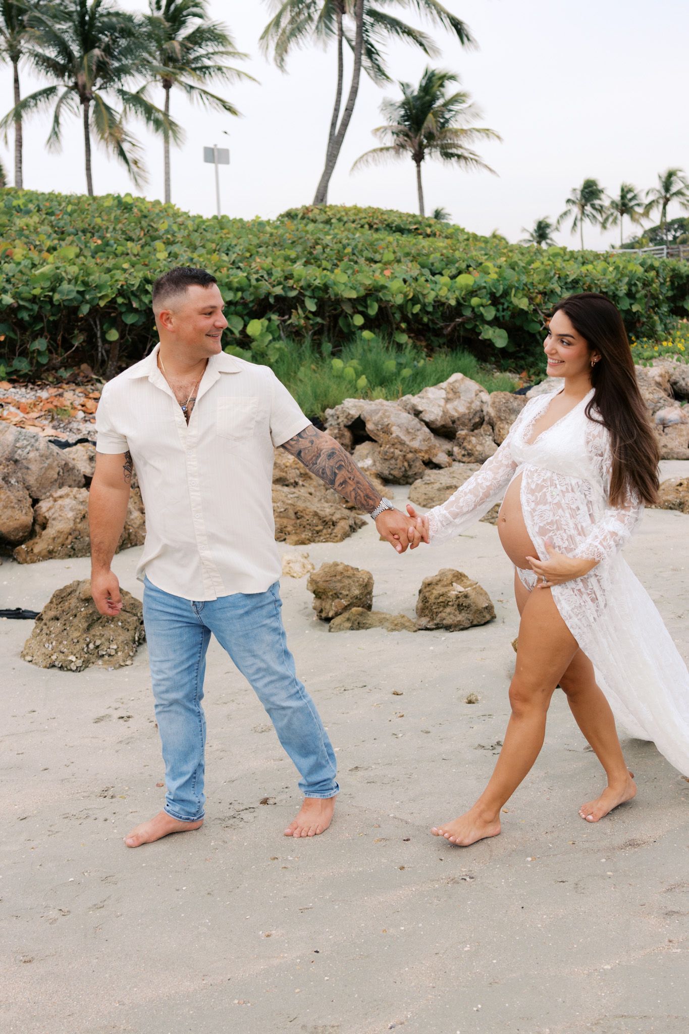 Couple holding hands and walking barefoot on a sandy beach with palm trees and rocks in the background.