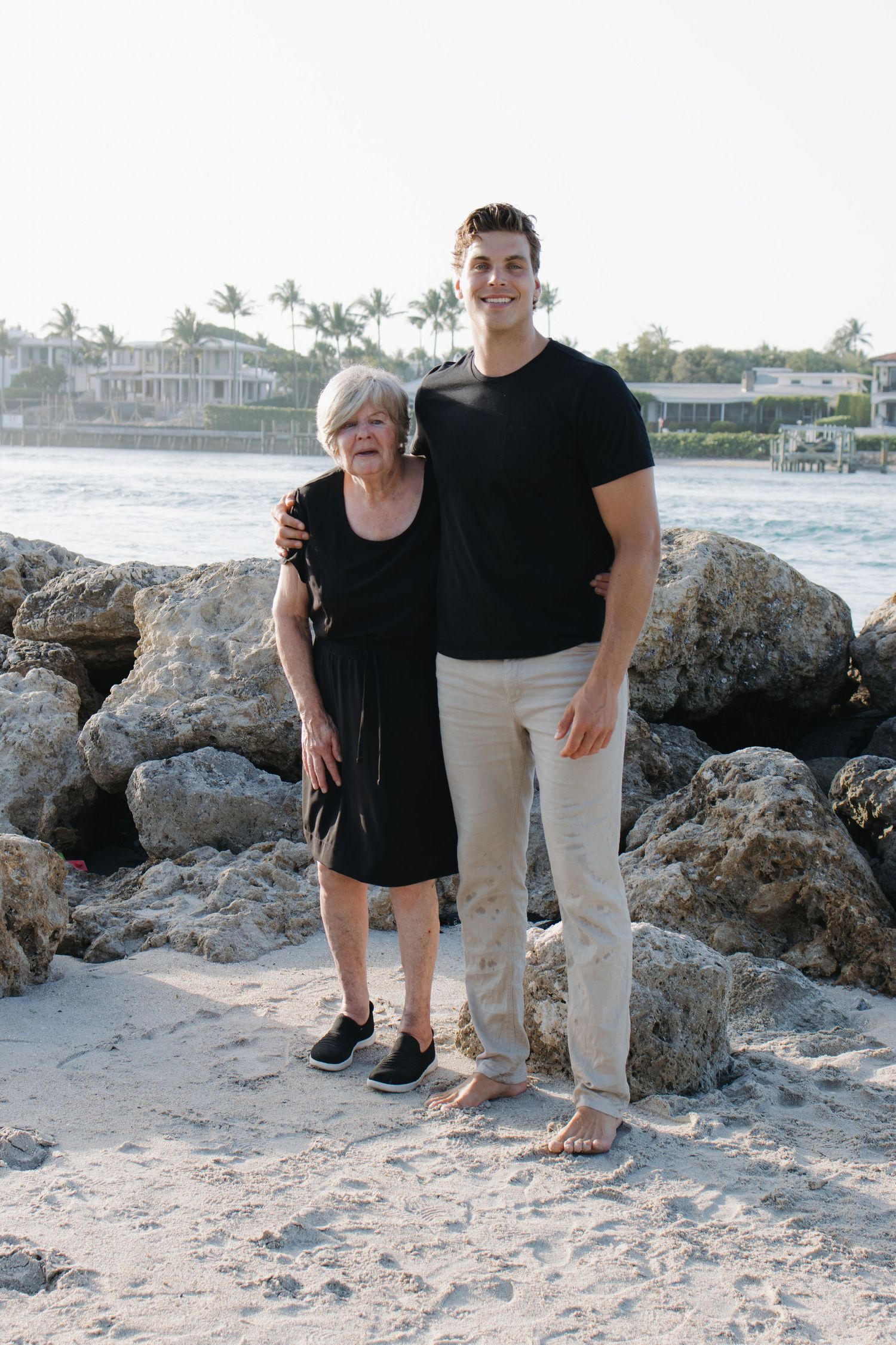Two people stand together on a rocky beach with luxury waterfront homes and palm trees in background.