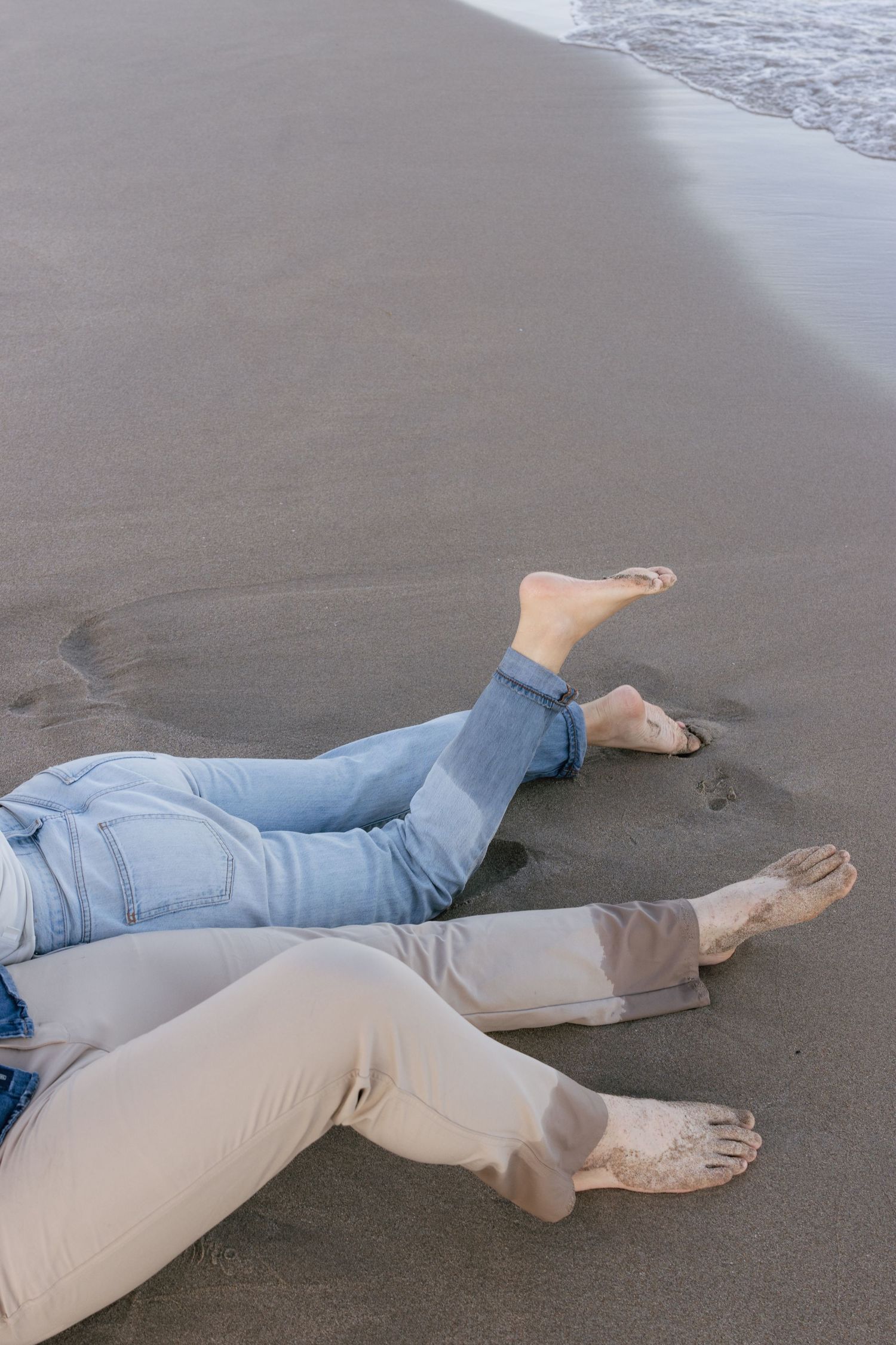 Couple lying on wet sand at beach wearing casual denim and white clothing.